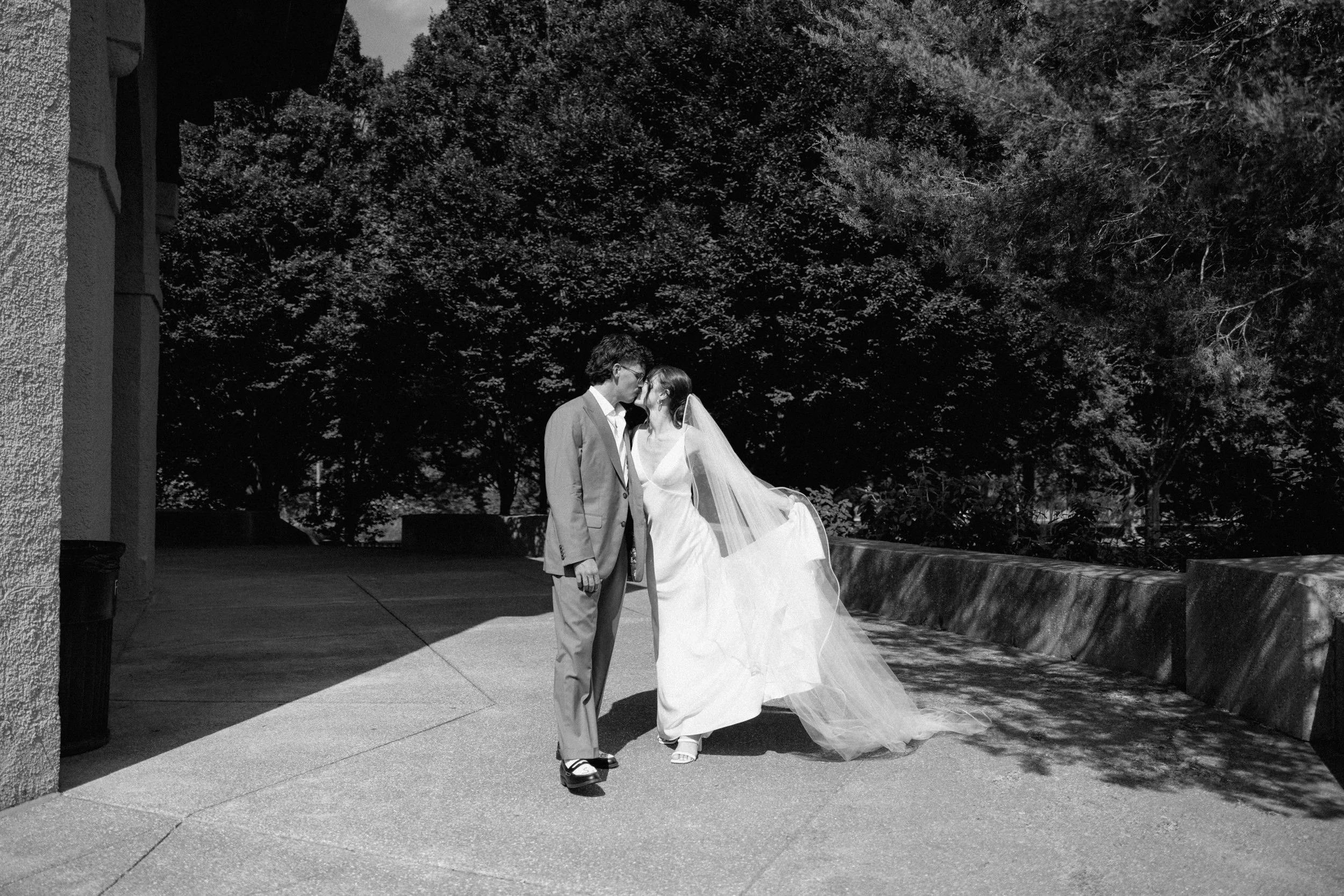 A black and white photo of a bride and groom standing outdoors, facing each other, with the bride twirling in her wedding dress while the groom looks at her, with large trees in the background.