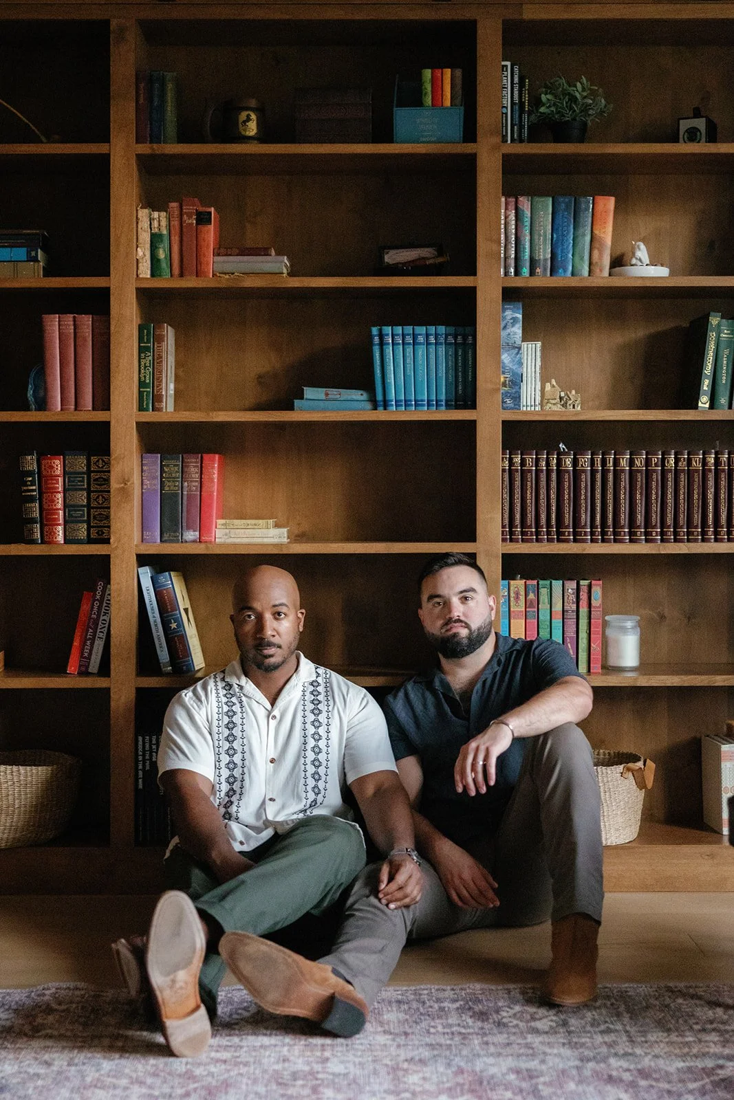Two men in dressed up attire holding hands and looking at the camera with a bookcase behind them