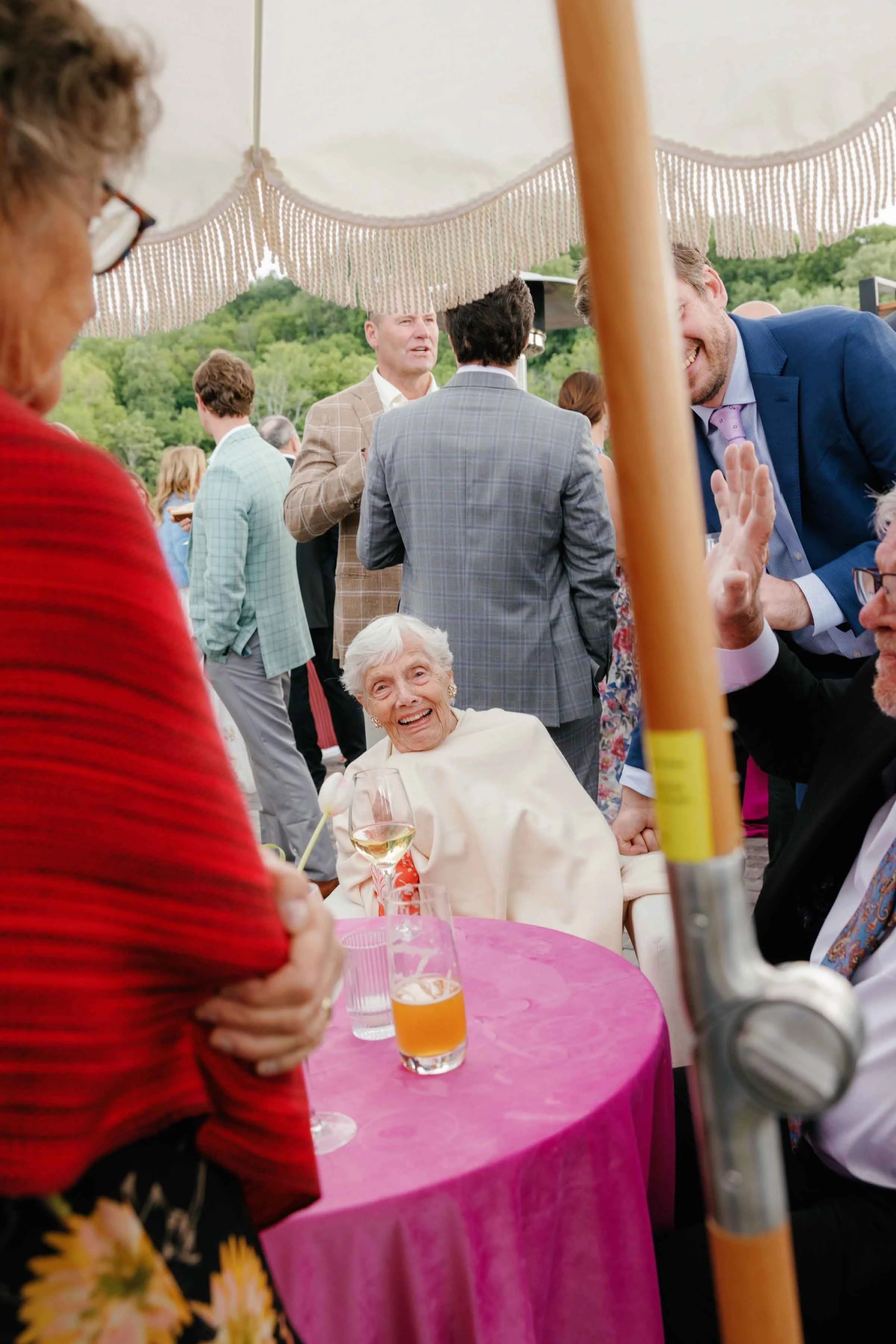 Older woman sitting at a pink table under an umbrella with other wedding guests smiling