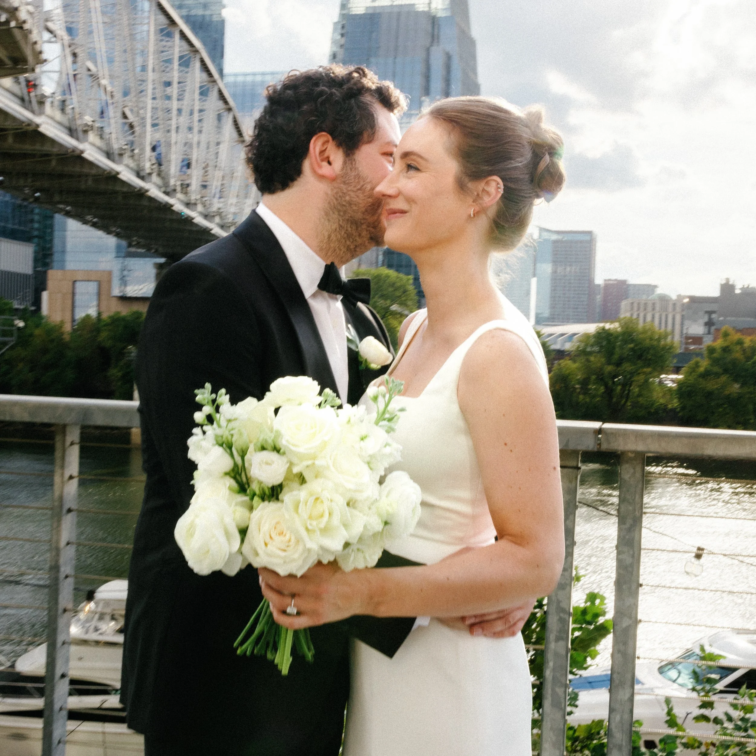 A man and woman sharing a close, intimate moment outdoors in front of the Nashville city skyline with a bridge in the background. The woman is holding a large bouquet of white flowers.