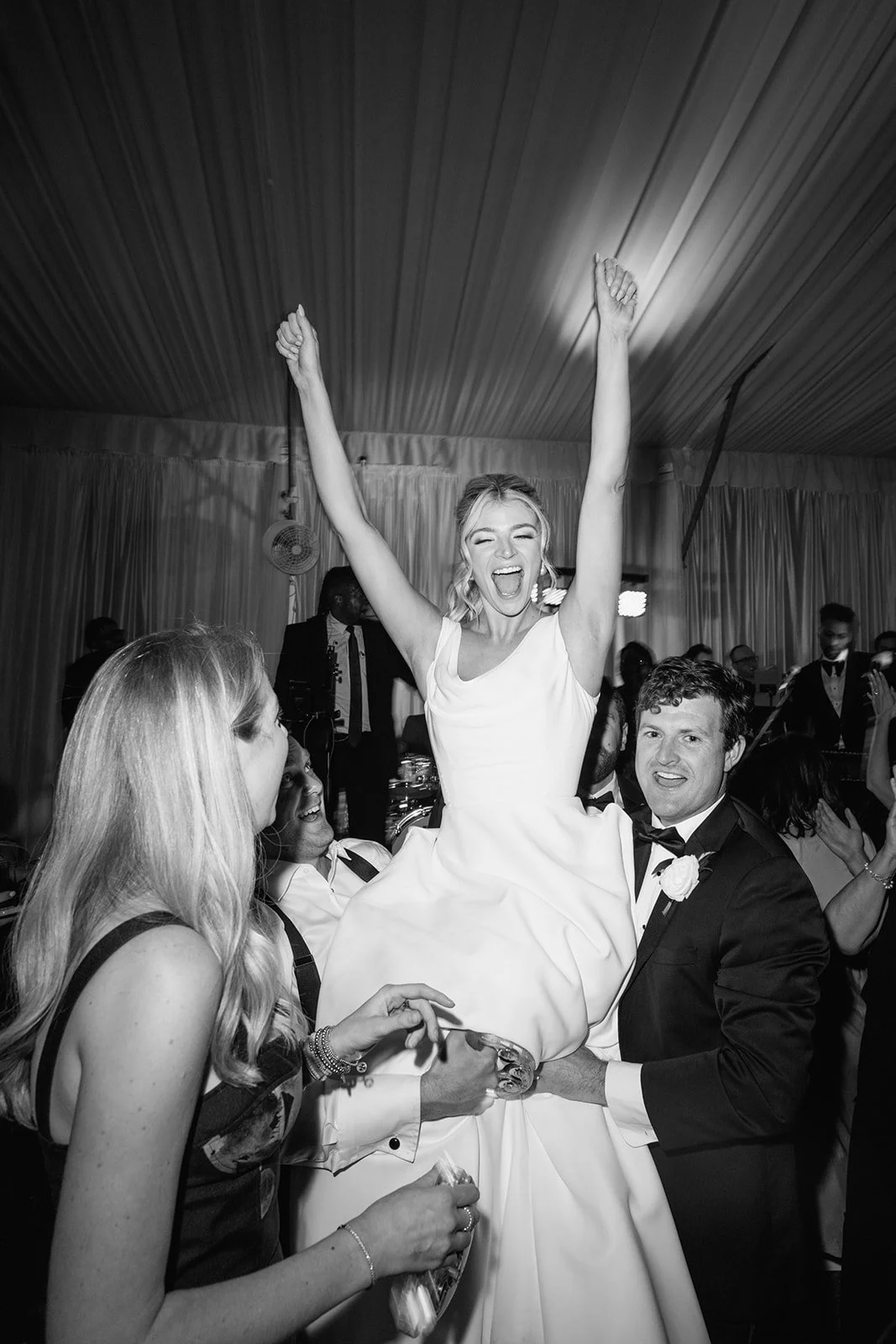 A joyful woman in a white dress is lifted in the air by wedding guests to celebrate. She has her arms raised in the air and is smiling widely. Other people are around her, smiling and cheering, in a decorated wedding venue space.