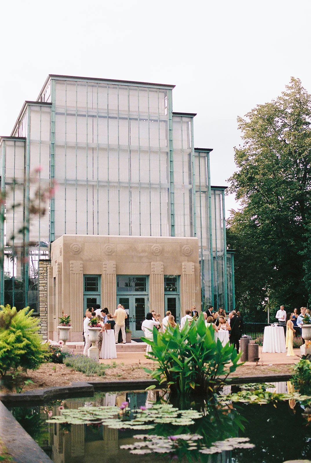People gather outside the Jewel Box in St. Louis, near a pond with lily pads and pink flowers, during a wedding.
