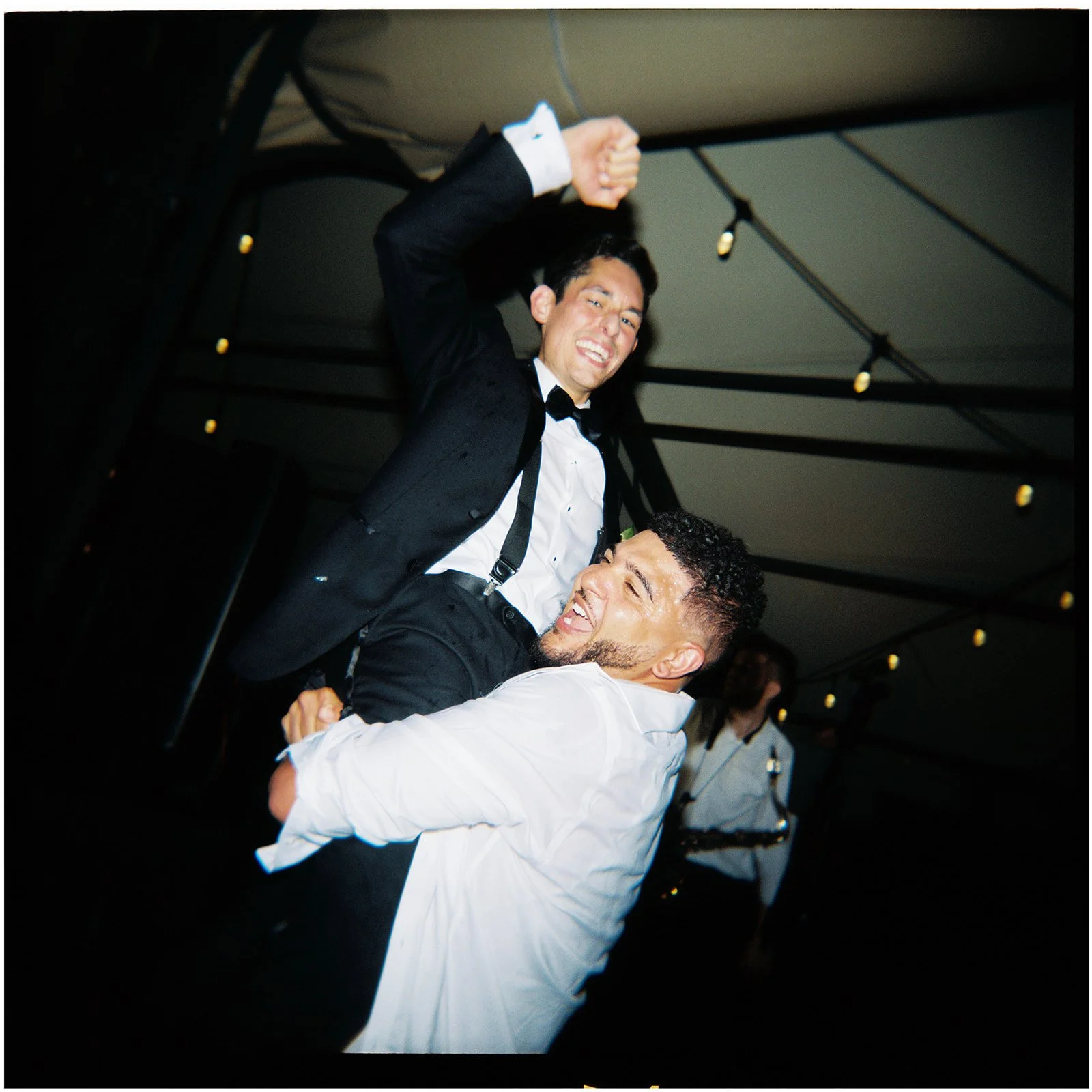 A film photo of two men in formal attire dancing closely at a party, one lifting the other in a dance move.