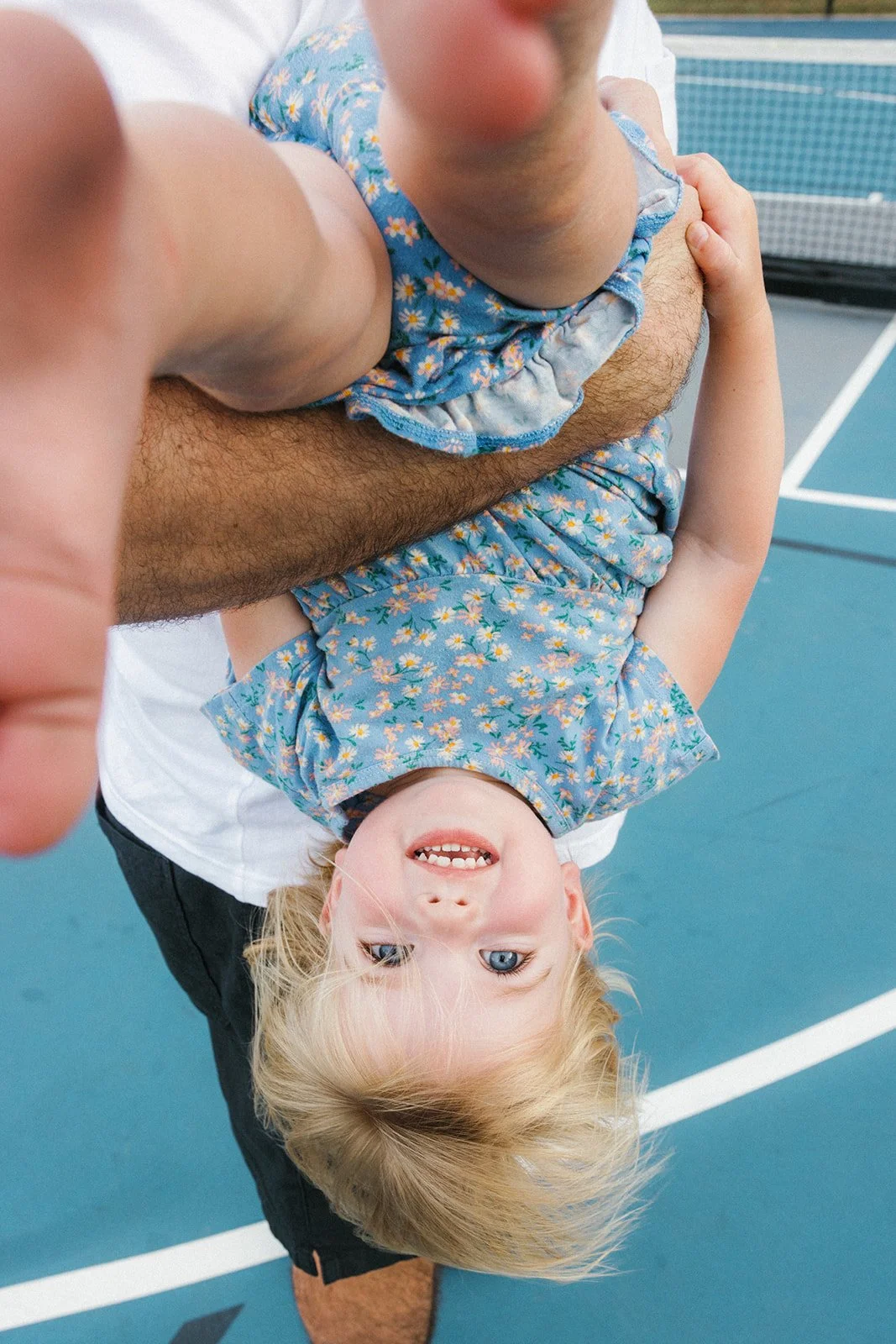 Kid behind held upside down in a playful way smiling on a tennis court in a blue outfit