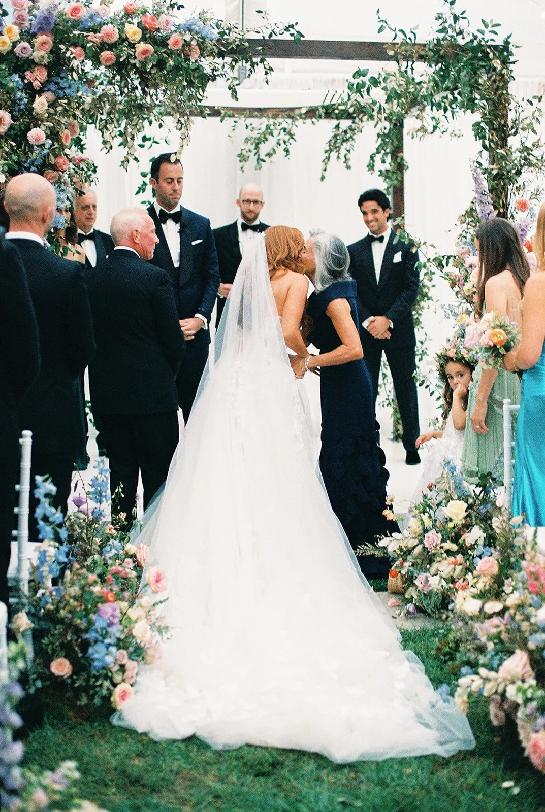 Bride and mom hugging at wedding ceremony. Bride in white dress with veil, groom in dark suit. Surrounded by wedding guests and floral decorations.