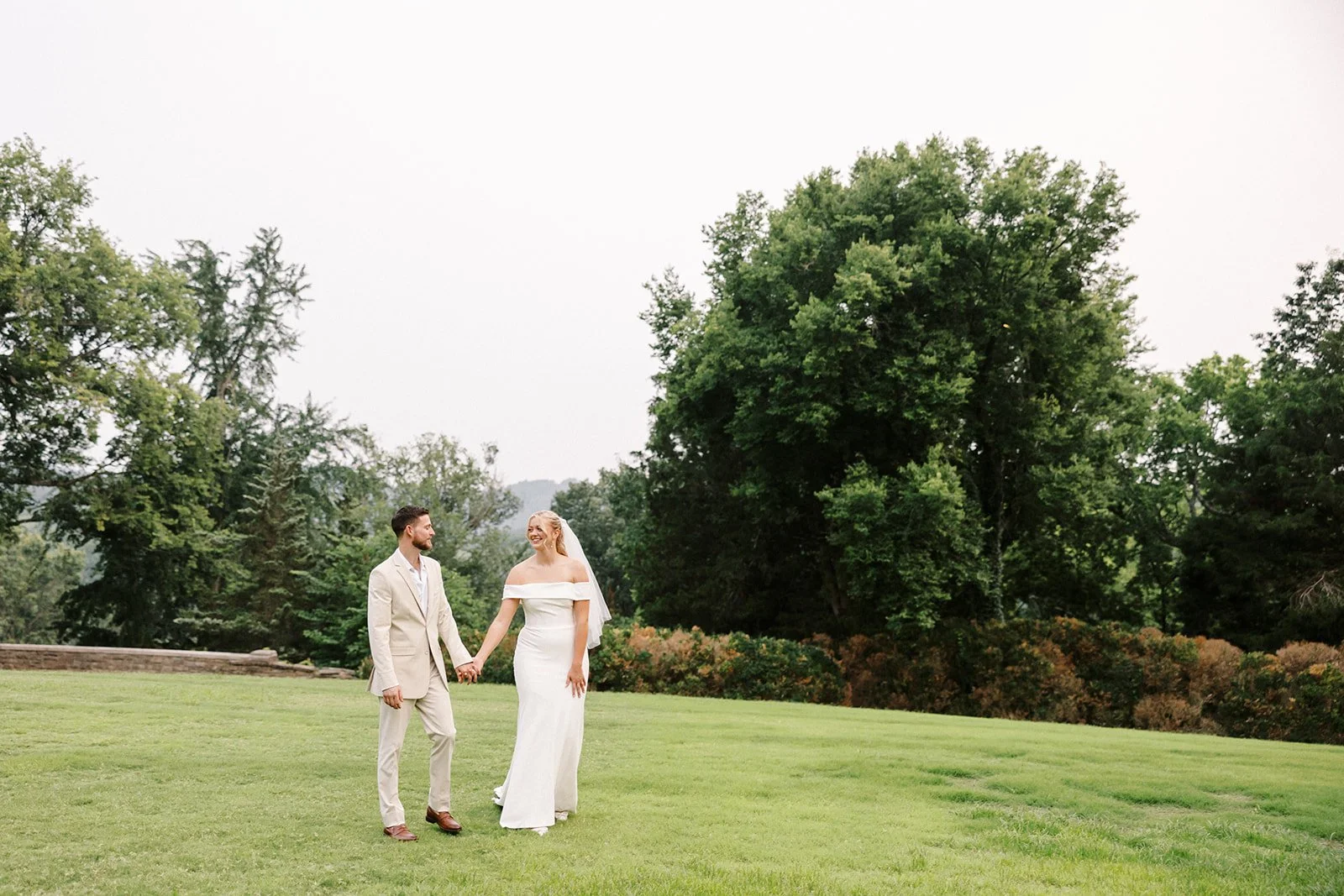 A bride and groom holding hands and walking on a grassy lawn during their outdoor wedding, with large trees and a clear sky in the background at Cheekwood Gardens.