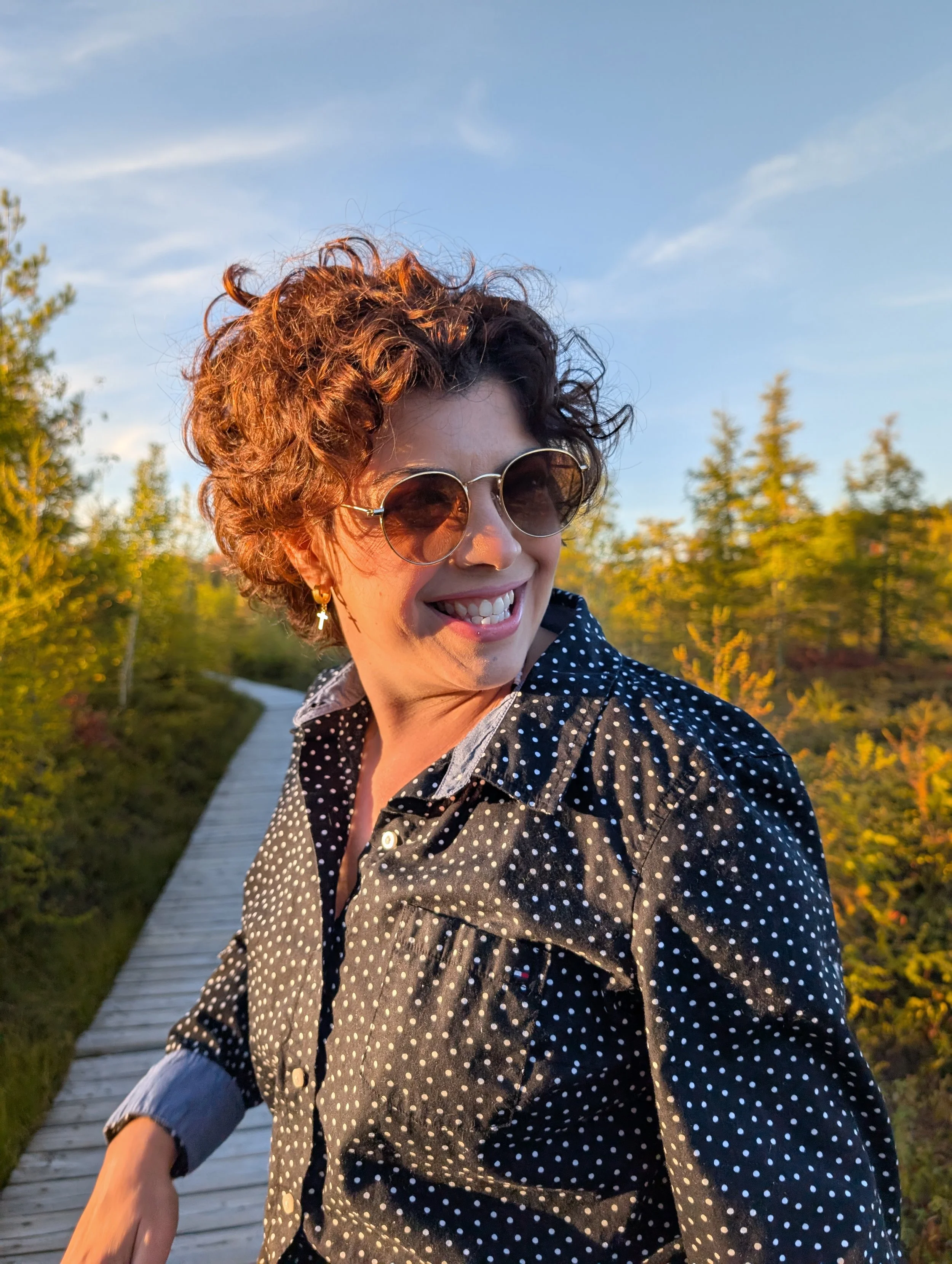 A woman with short curly hair and wearing sunglasses, surrounded by green trees during sunset.