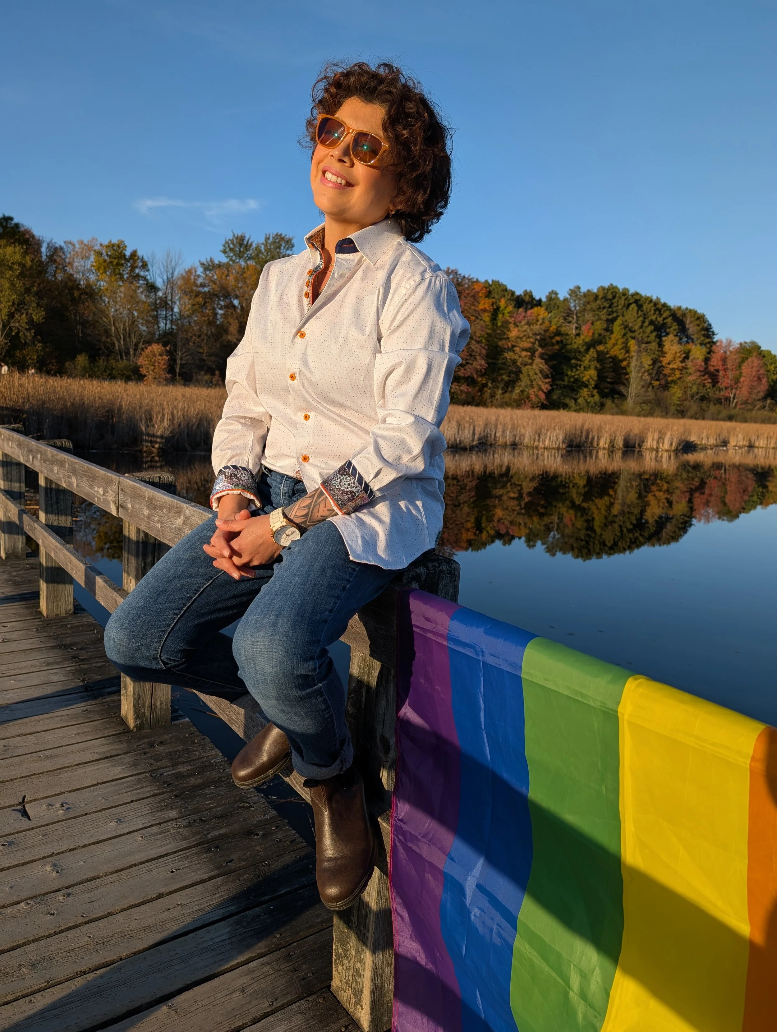 A woman with curly hair, sitting on a wooden dock next to a rainbow pride flag, with a lake and trees in the background.