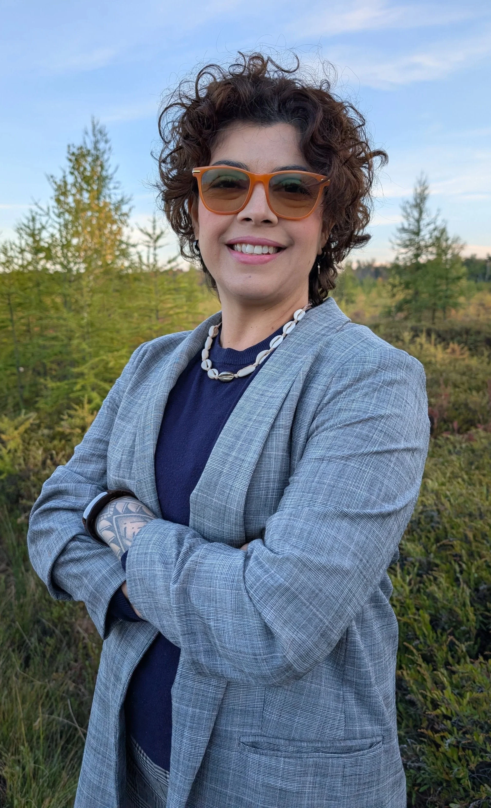 A woman with short curly hair wearing sunglasses with a background of green trees.