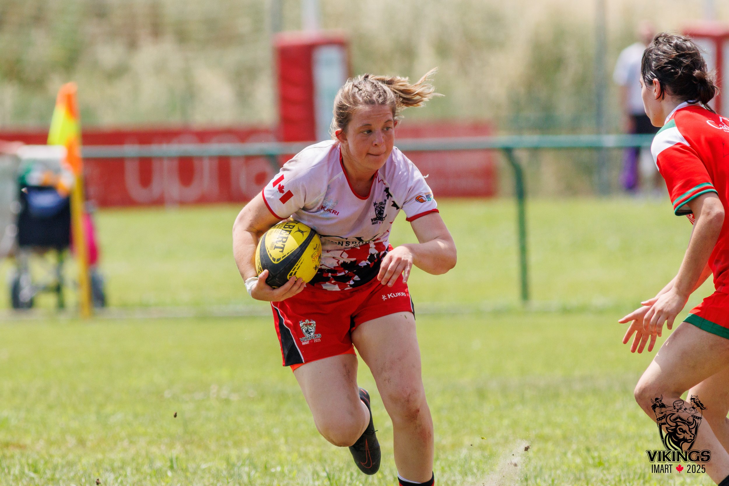 A female rugby player in a white and red jersey carrying a yellow rugby ball running on a grassy field during a match.