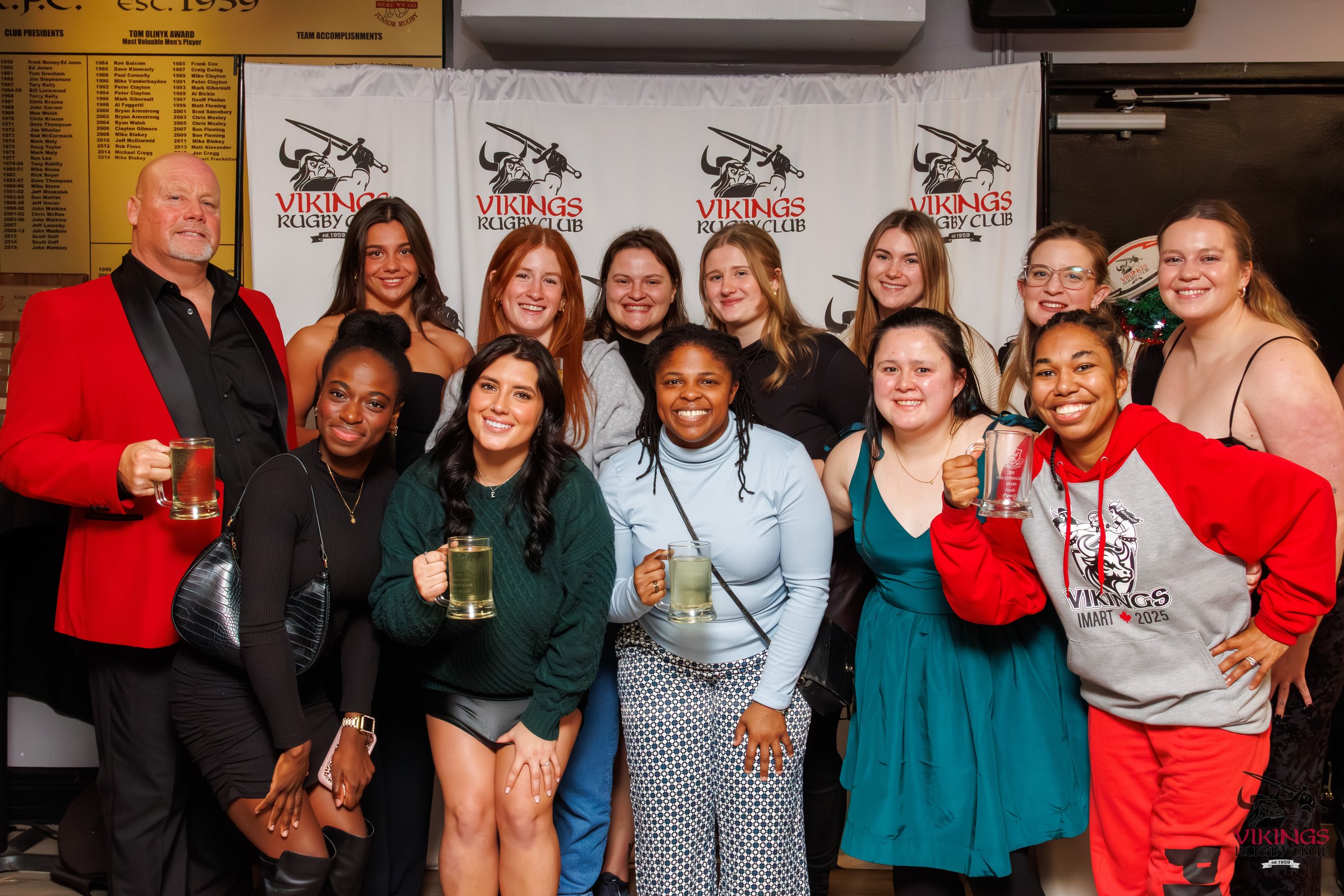 A group of people at a Viking rugby club event, posing with drinks, in front of a Viking Rugby Club backdrop, smiling and celebrating.