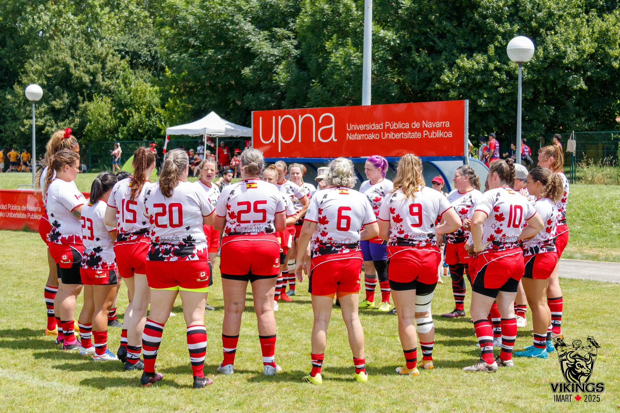Women rugby team wearing white jerseys with red and black designs, red shorts and striped socks, standing in a circle on a grassy field during daytime, with the UPNA university sign and other players and spectators in the background.