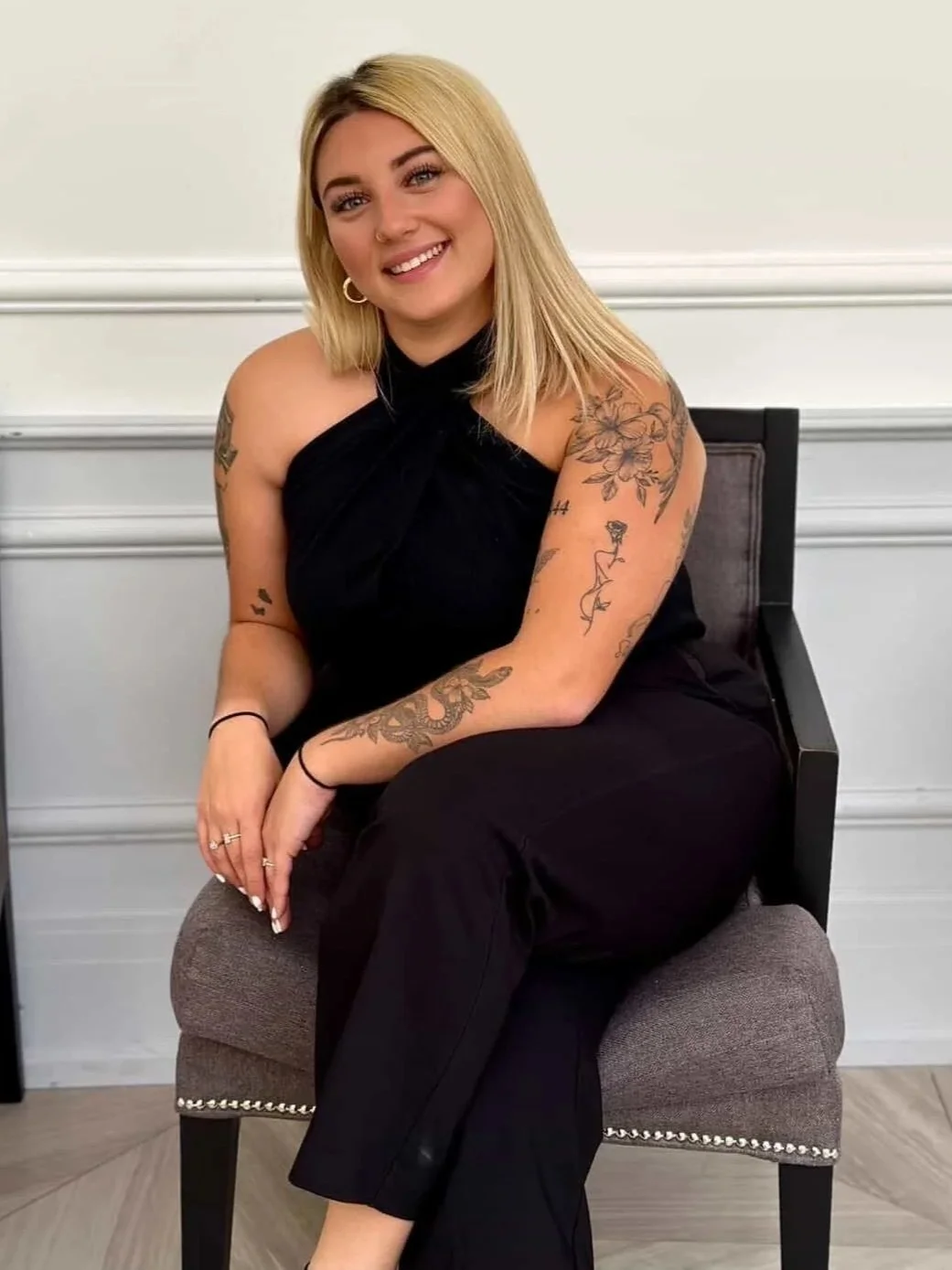 A smiling woman with blonde hair, wearing a black halter top and pants, sitting on a chair with a gray cushion, in front of a white wall with decorative molding.