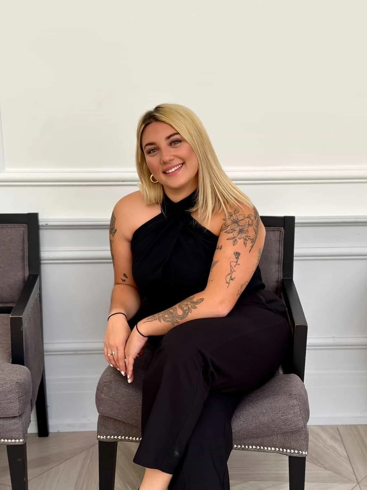A smiling woman with blonde hair, tattoos on her arms, wearing a black sleeveless top, sitting on a chair in front of a white wall.