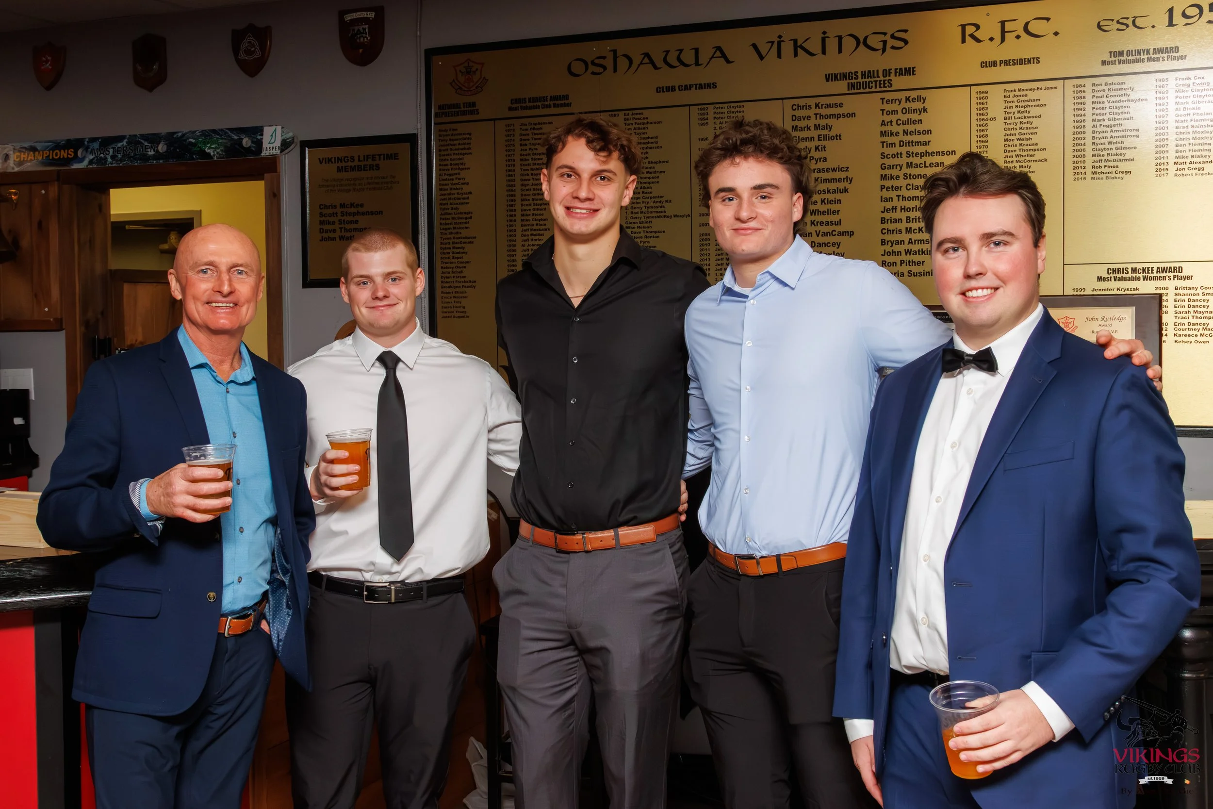 Group of five men dressed in formal attire, standing together indoors at a celebration or event, with a wall display in the background listing the Oshawa Vikings Rugby Club hall of fame inductees.