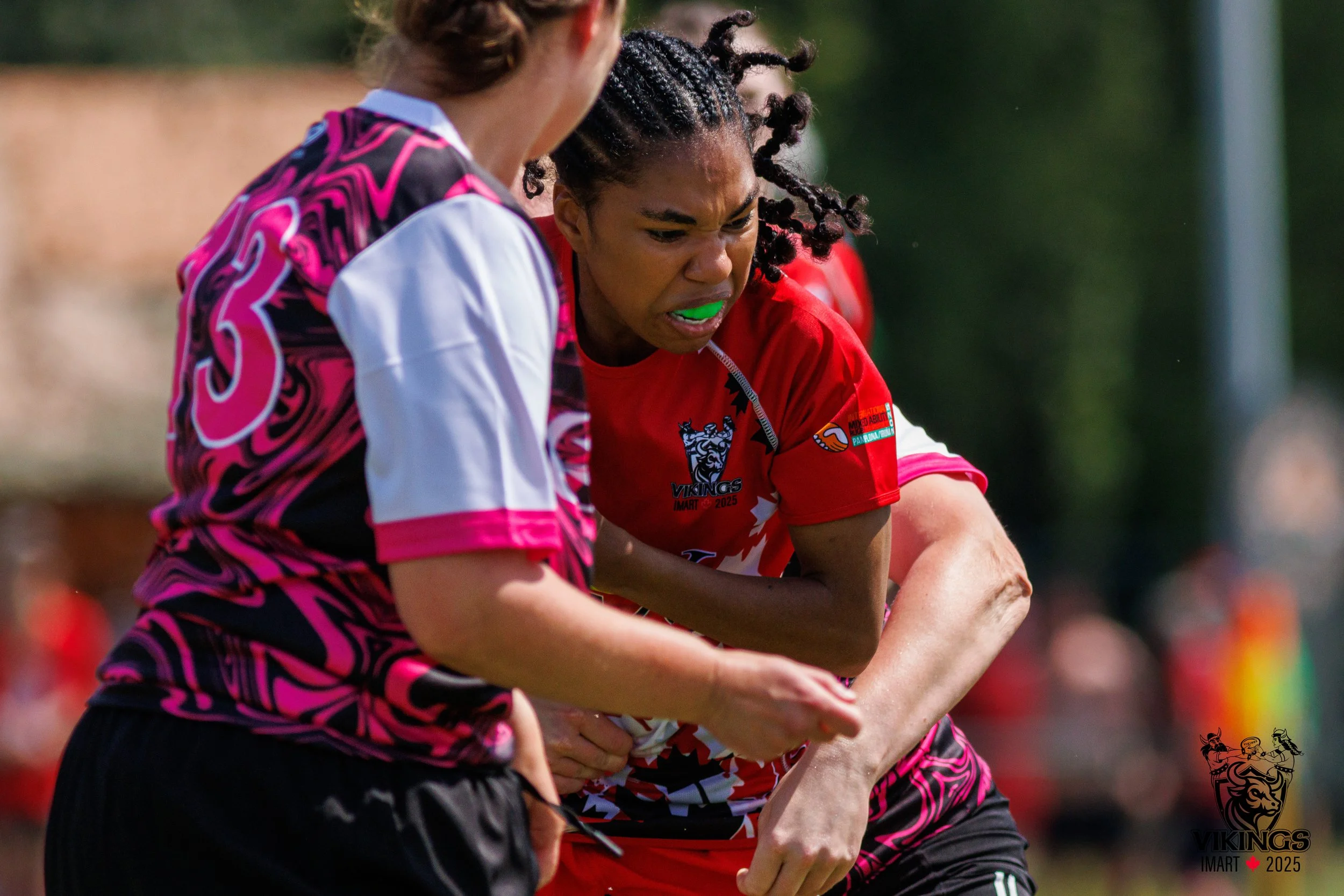 A female rugby player with braided hair wearing a red uniform grimaces with a mouthguard during a game, while an opponent in a black and pink jersey is in close proximity on the field.