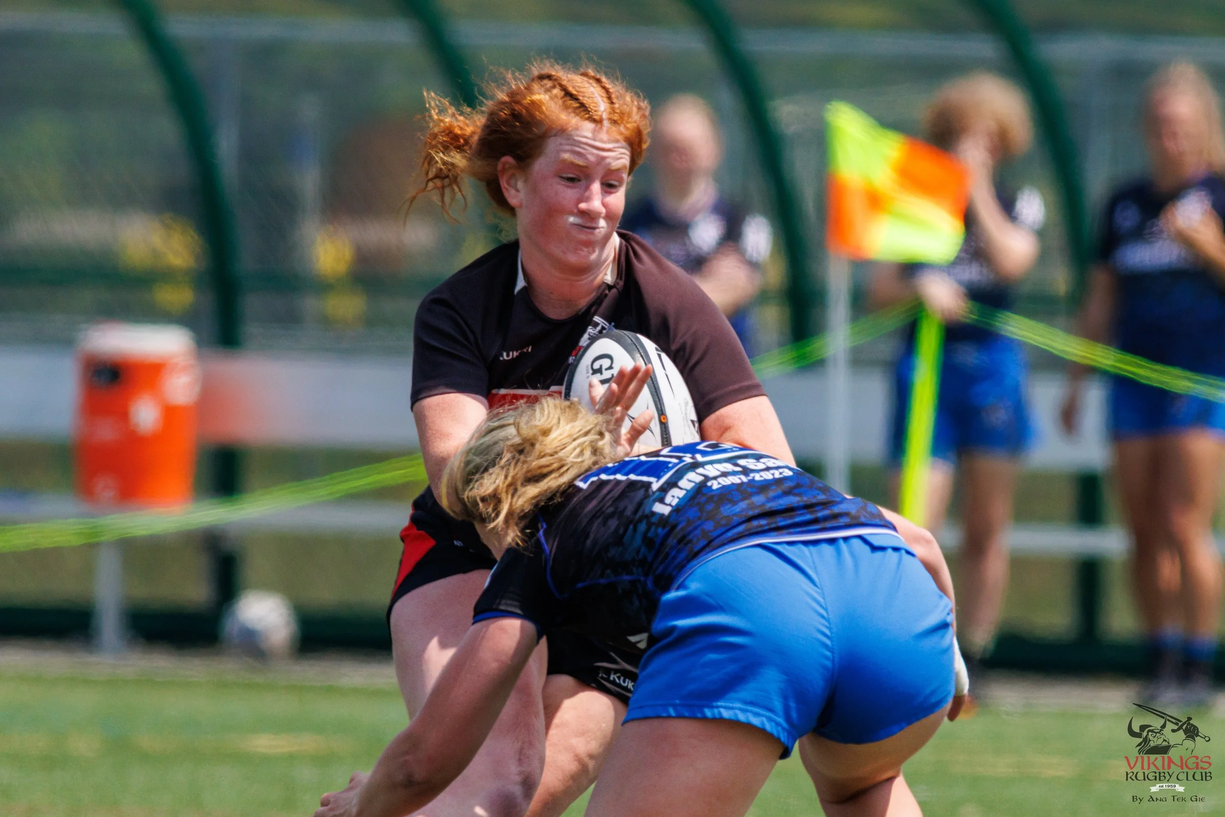 Women playing rugby, one in black jersey holding a rugby ball, another in blue shorts bent over, with others in the background near the sideline.