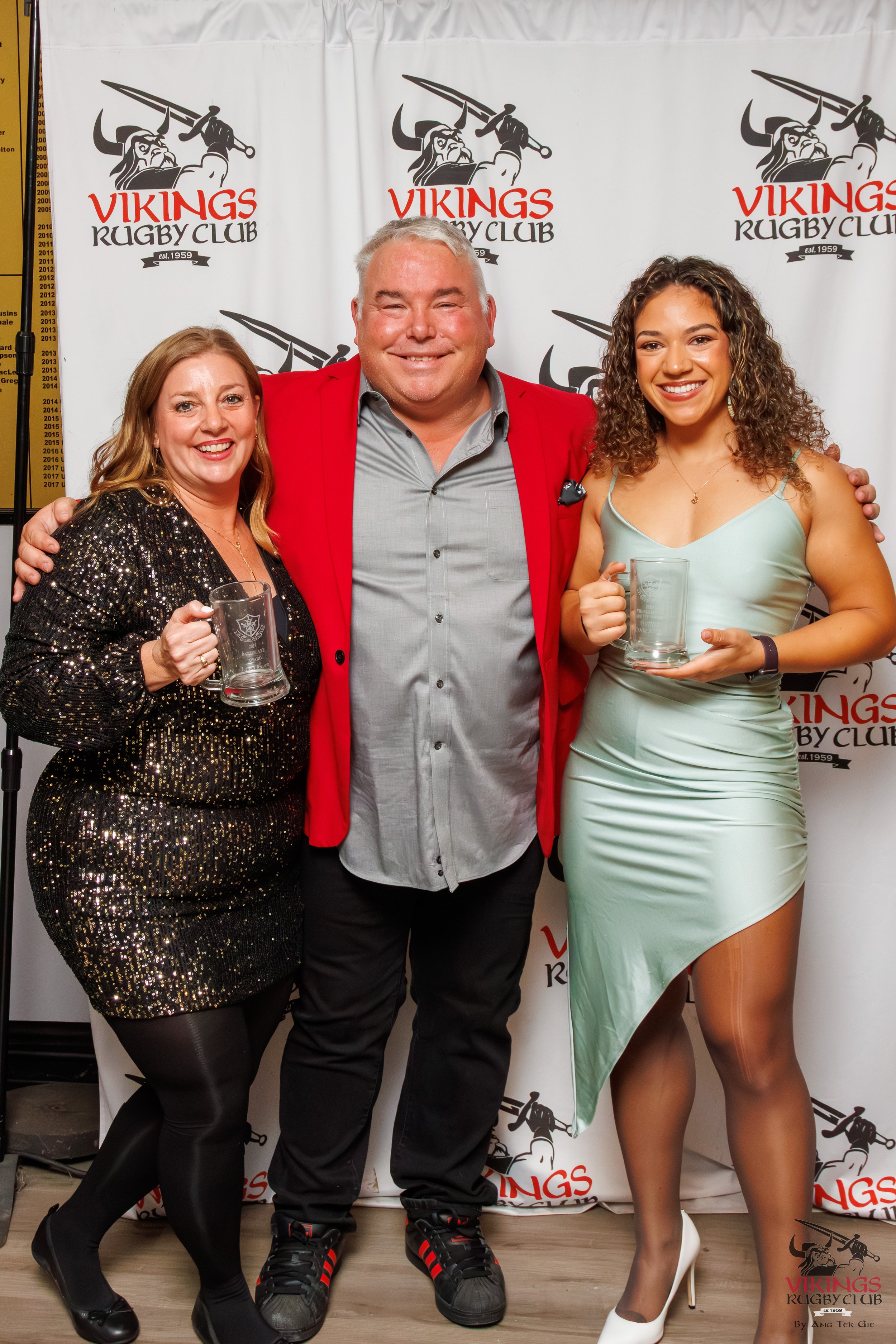 Three people smiling at Vikings Rugby Club event, holding beer mugs, standing in front of a white backdrop with the club's logo. The woman on the left wears a black glittery dress and black tights. The man in the center wears a gray shirt, red blazer