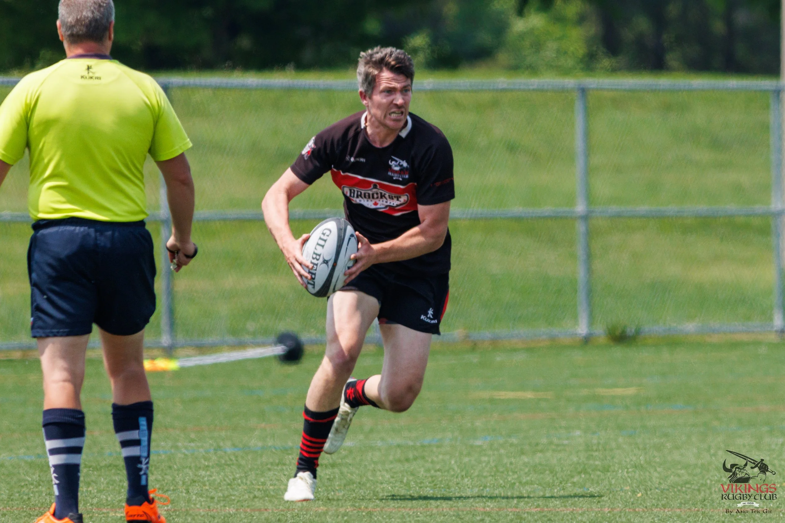 Rugby player running with ball during a game, referee observing, on field with fence in background.