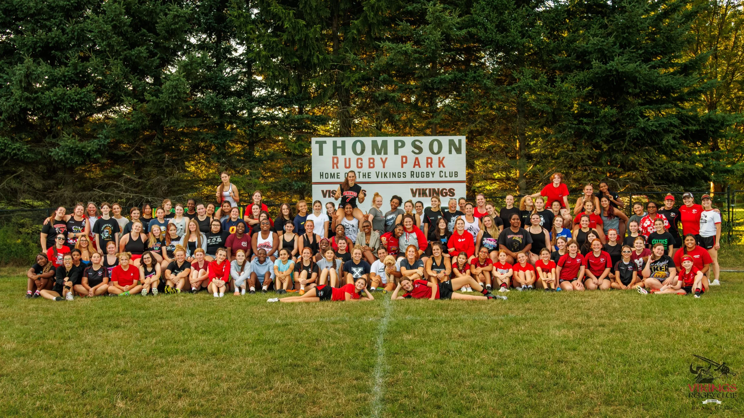 Large group of people, mostly young women, gathered on a grassy field in front of a sign that reads 'Thompson Rugby Park, Home of the Vikings Rugby Club.' They are posing for a group photo, many wearing sports jerseys and casual clothing, with some s