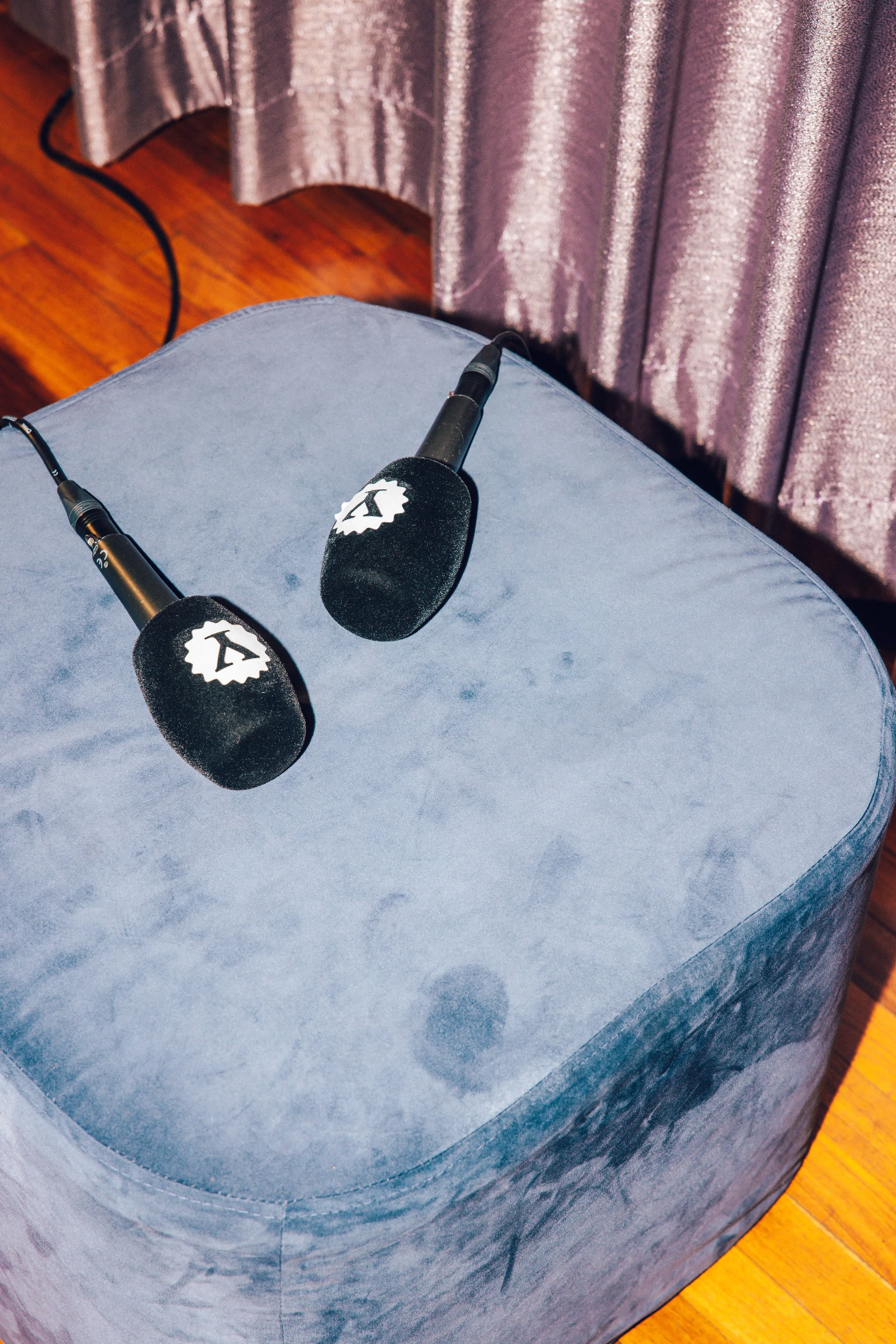 Two black microphones with foam covers featuring a white logo, resting on a blue plush ottoman, with pink curtains in the background.