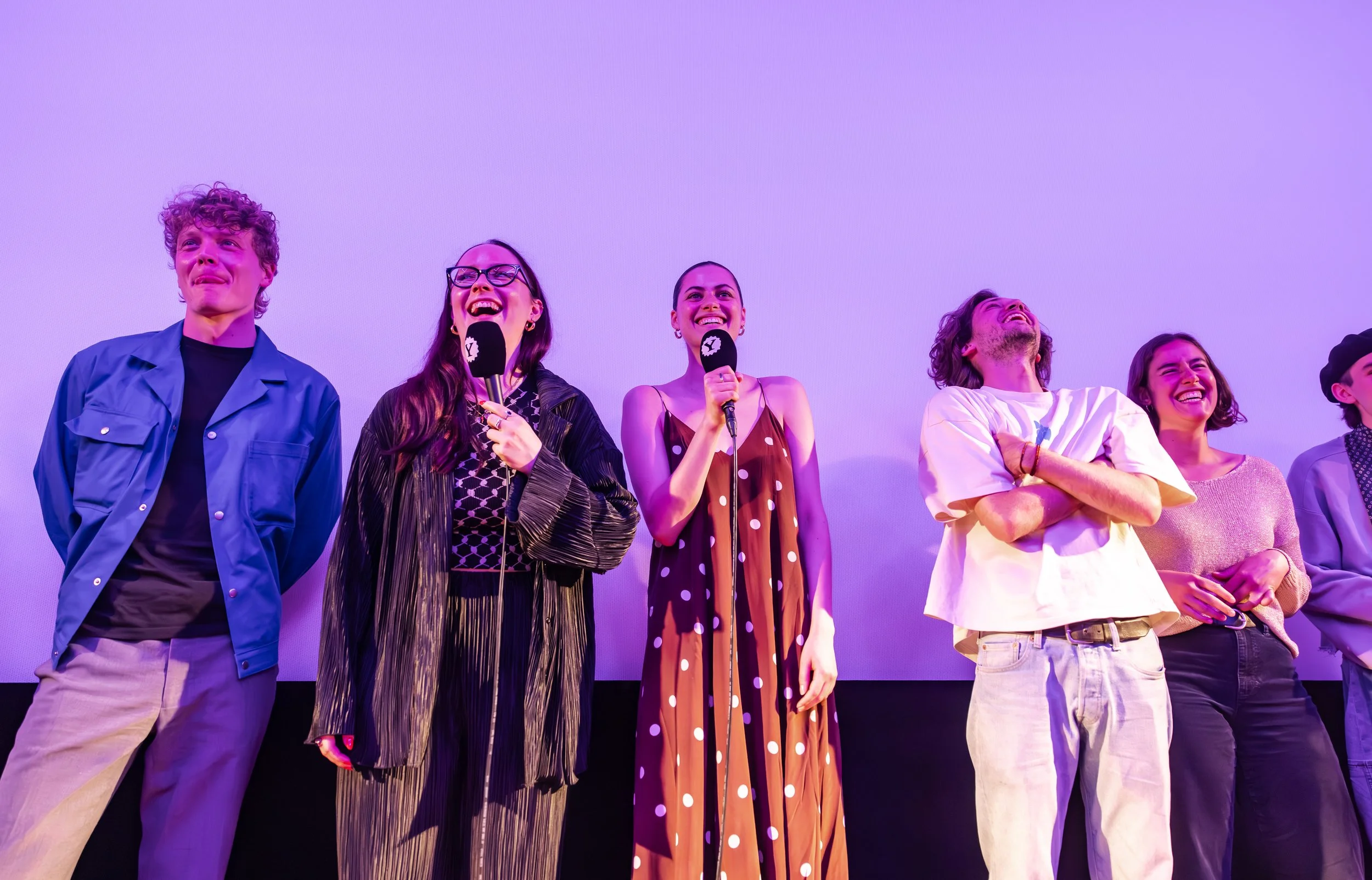 Group of six people standing on stage with a purple backdrop, some holding microphones, all smiling or laughing.