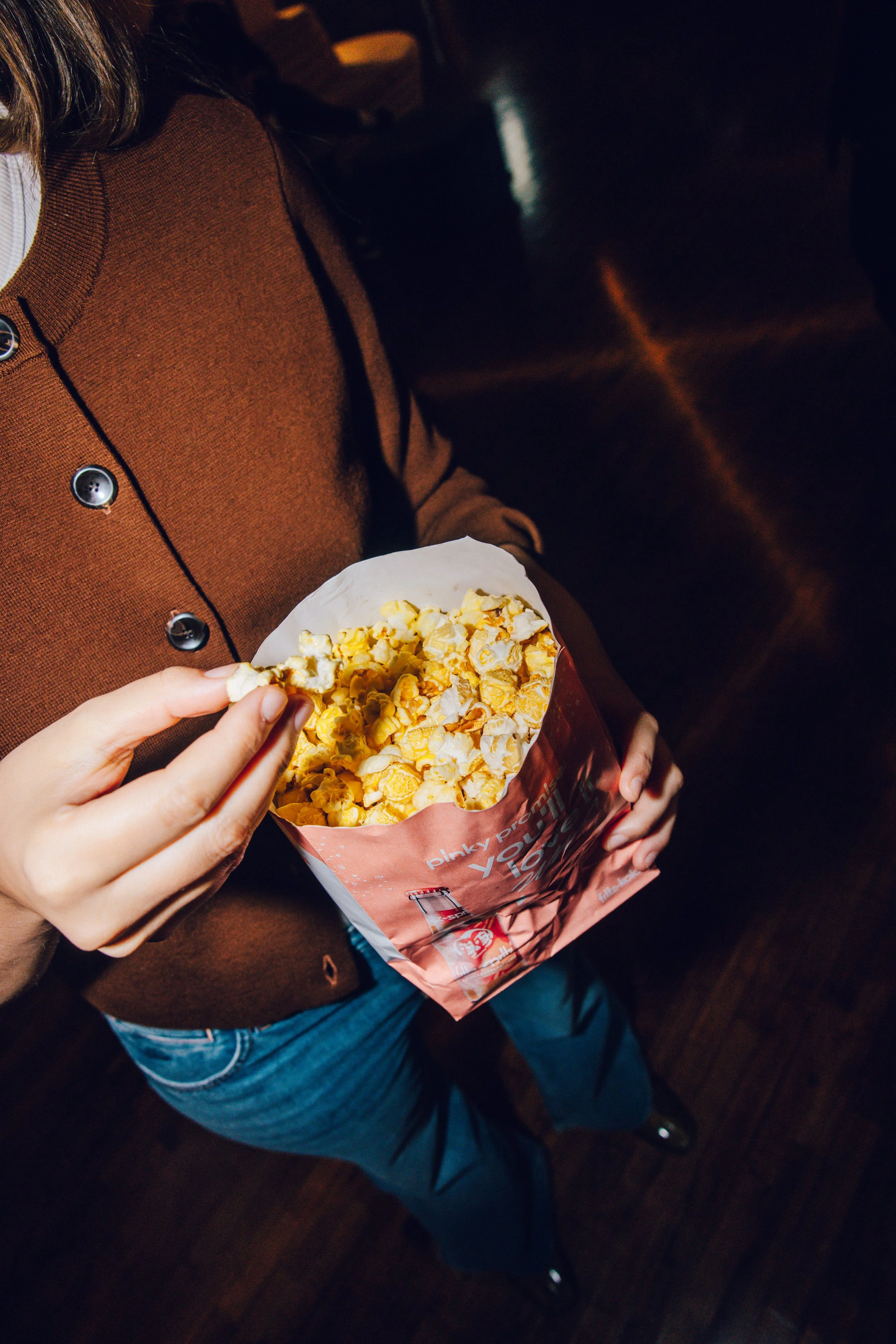Person holding a red and white bucket of buttered popcorn in a movie theater.