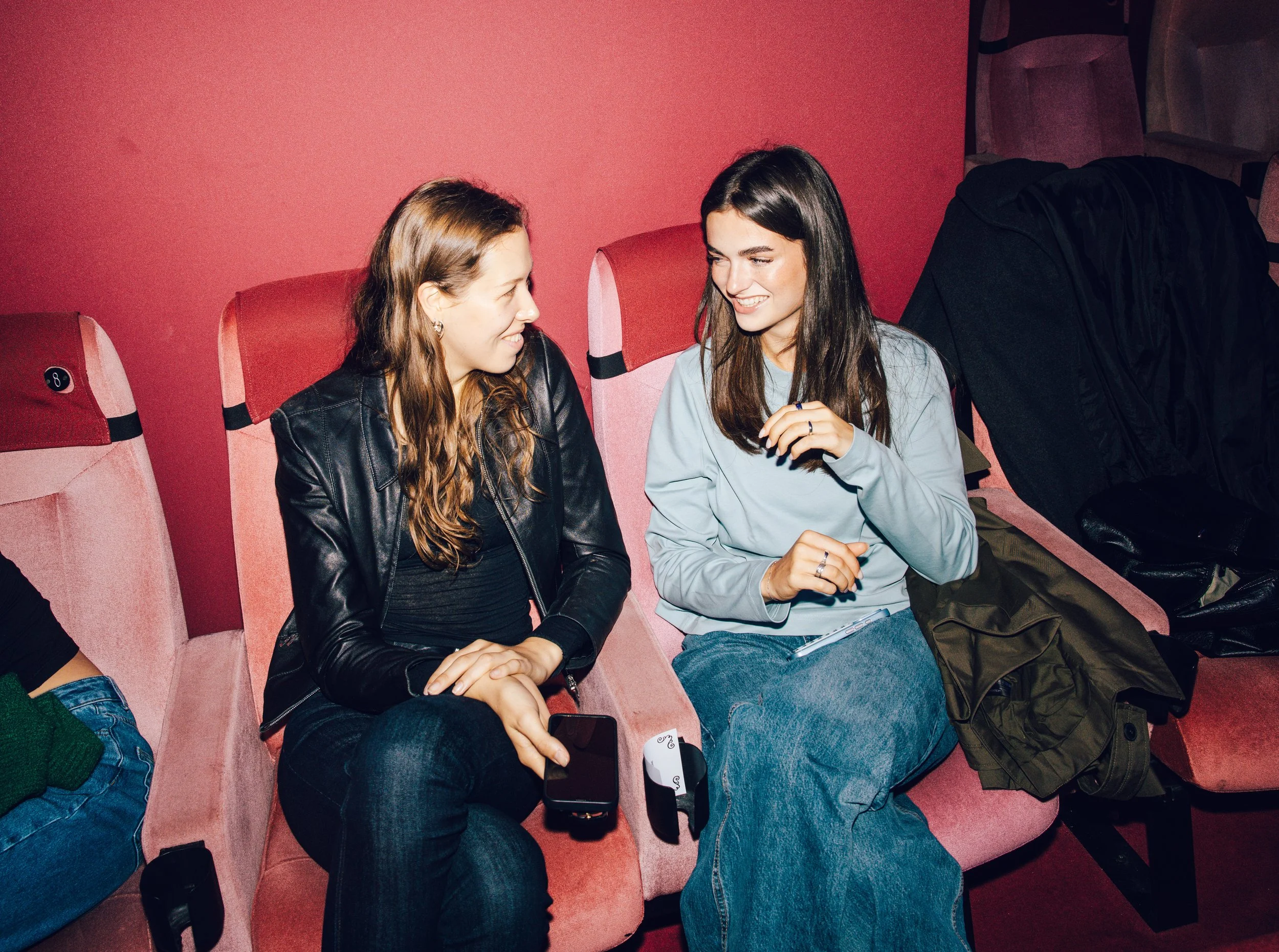 Two women sitting on pink theater seats, chatting and smiling at each other. One woman has long brown hair, wearing a black leather jacket and black pants, holding a smartphone. The other woman has long dark hair, wearing a light blue sweatshirt and 