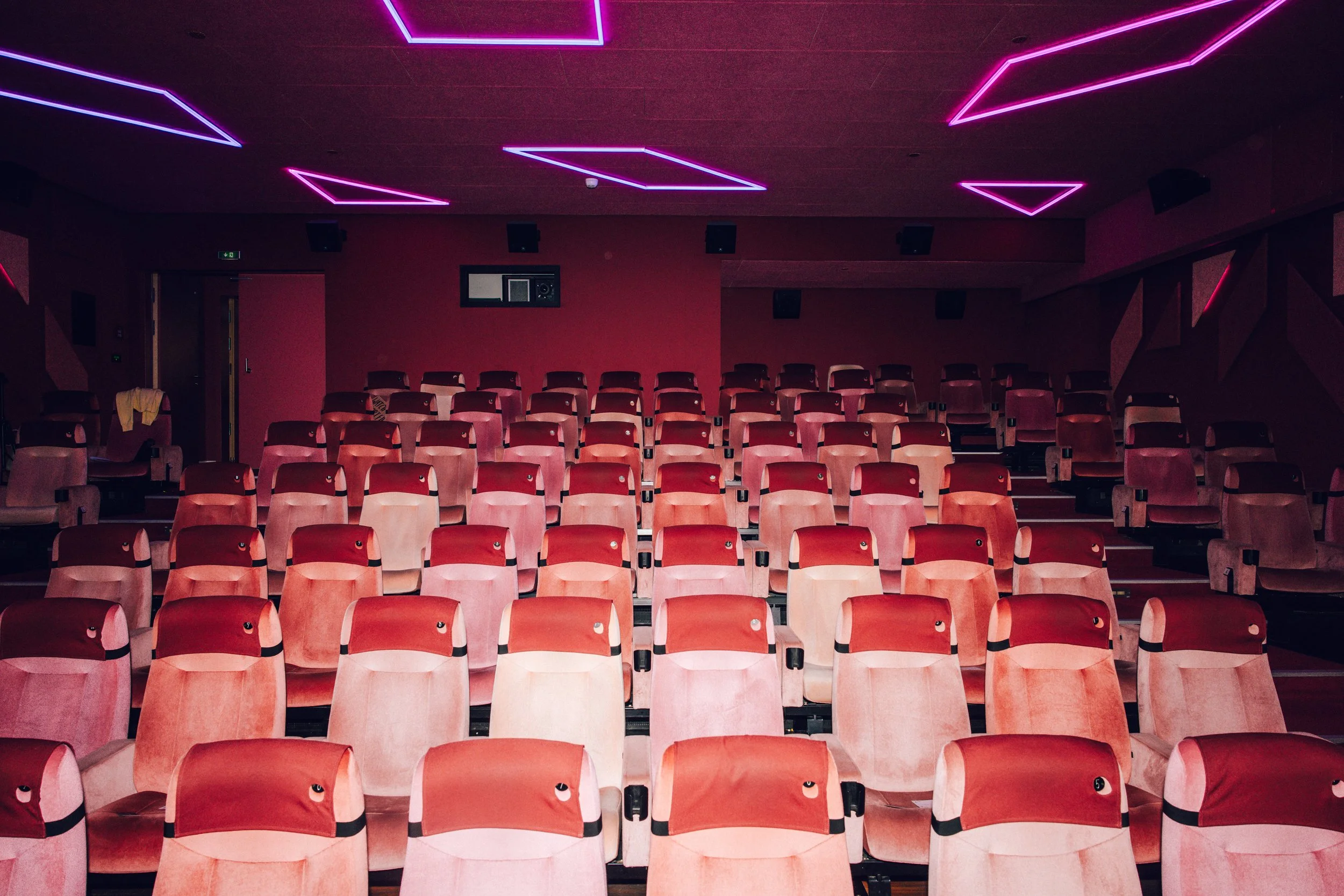 An empty movie theater auditorium with rows of red cushioned seats and purple neon lighting on the ceiling.