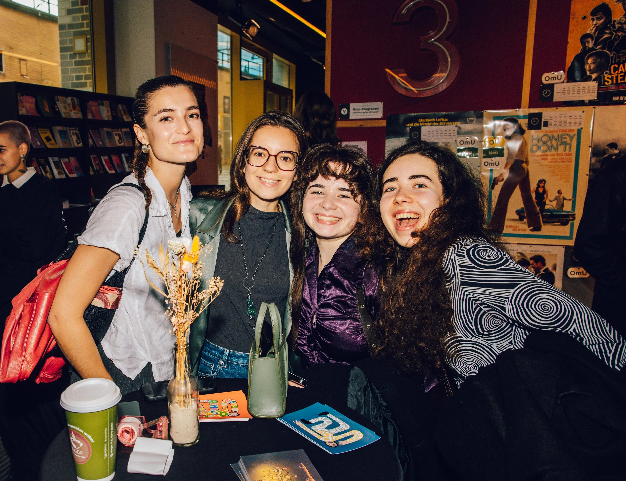 Four young women smiling and posing for a photo at a movie theater or cinema lobby, with posters and a large number 3 in the background.