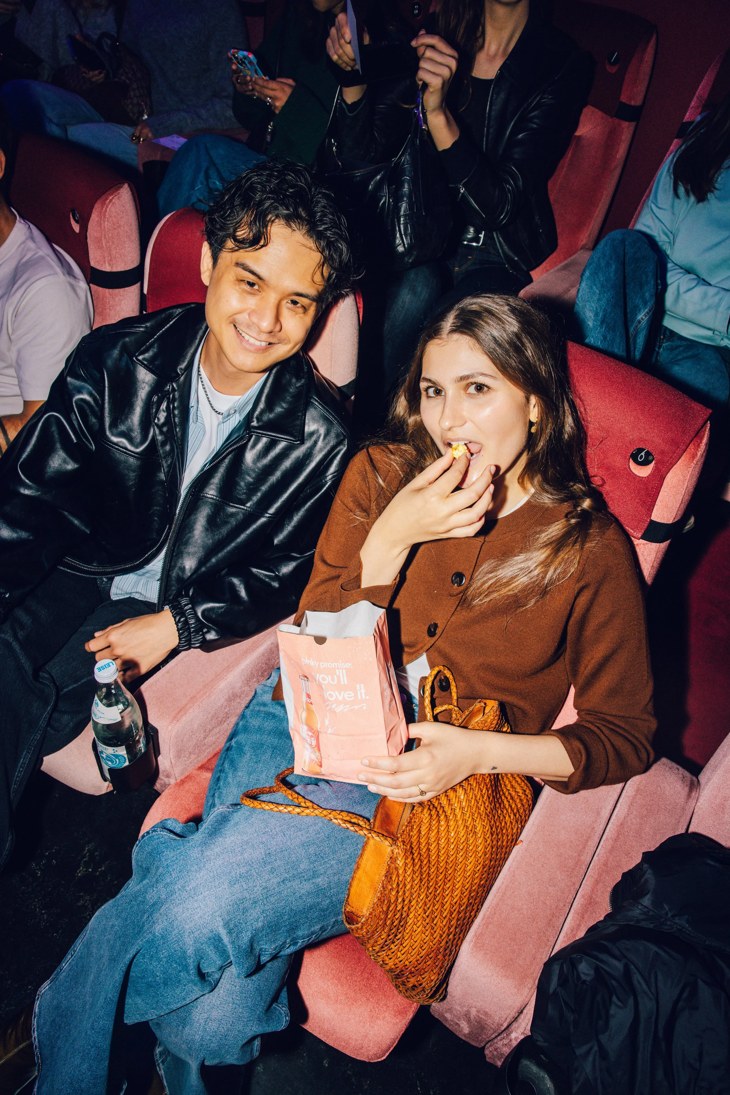 A young man and woman sitting in a movie theater, with the woman eating popcorn.