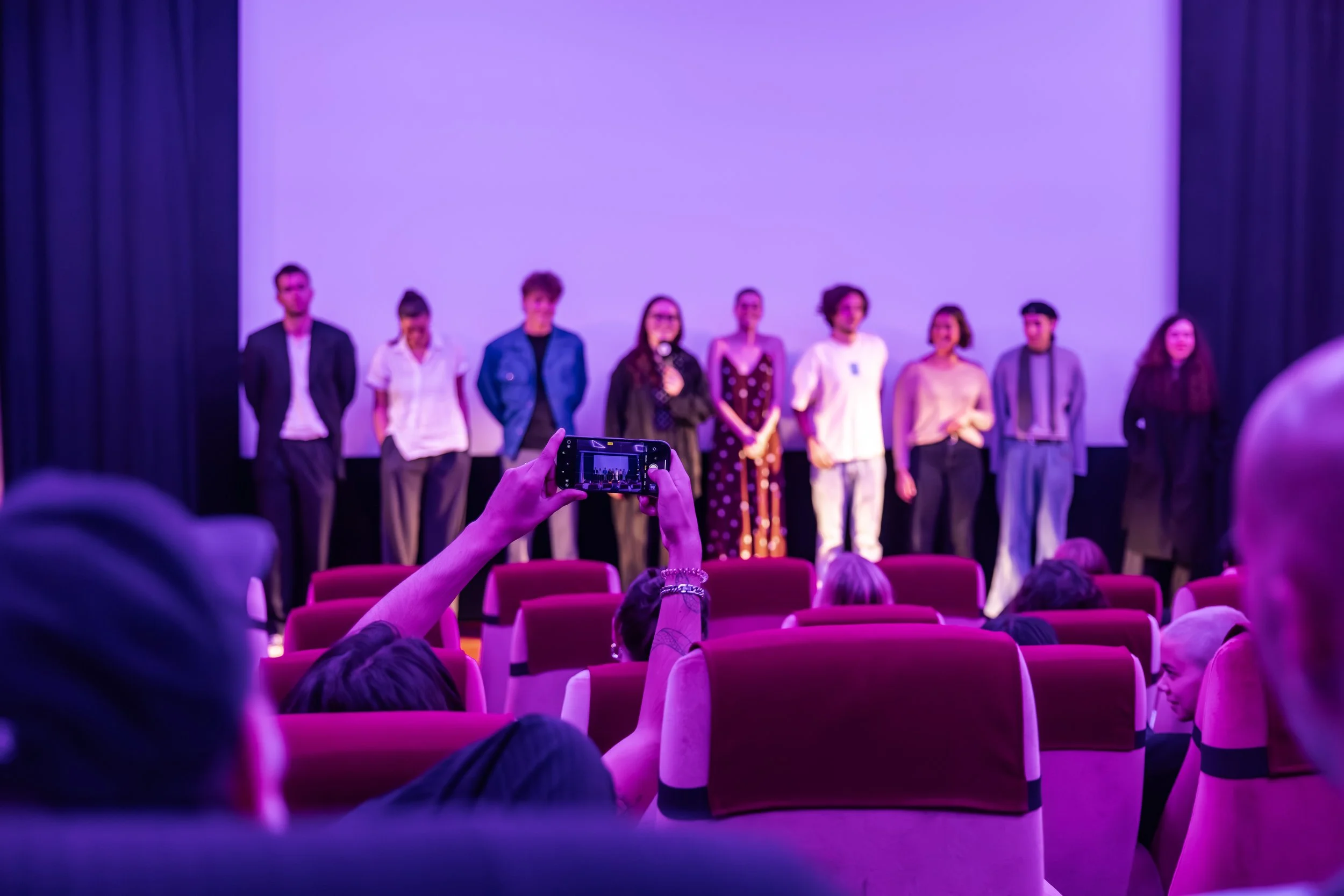 Group of people standing on stage in front of a large screen during a presentation or event, with an audience seated in front and someone in the audience taking a photo.