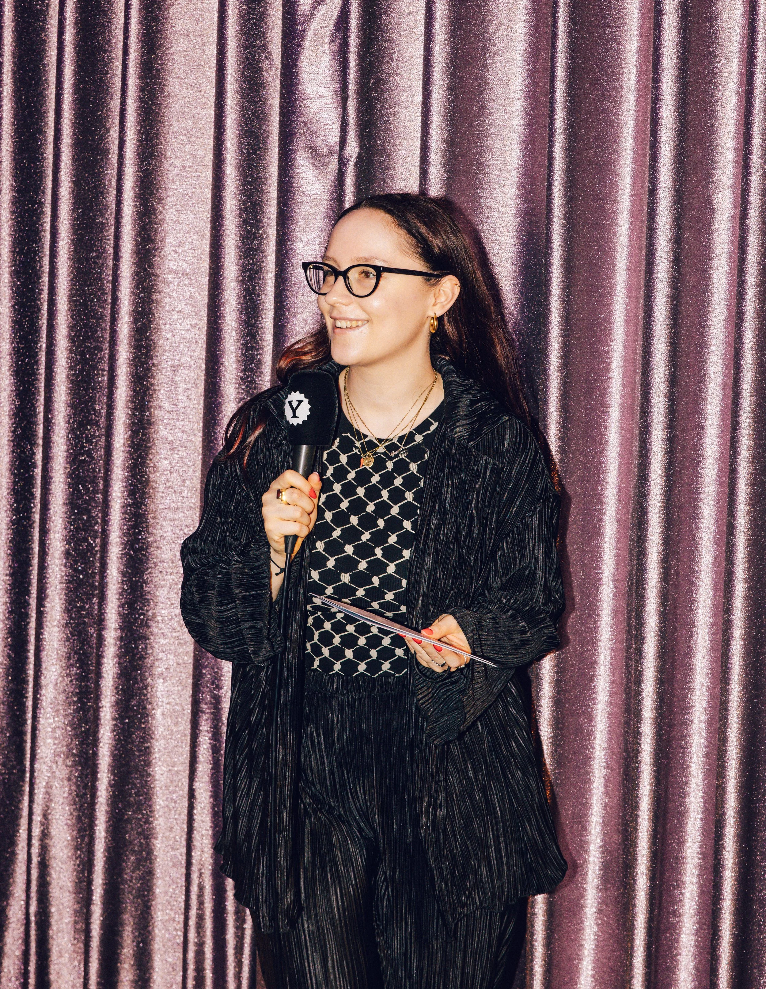 A woman with glasses and long hair, dressed in black patterned clothing, holding a microphone and a notepad, standing in front of a pink curtain.