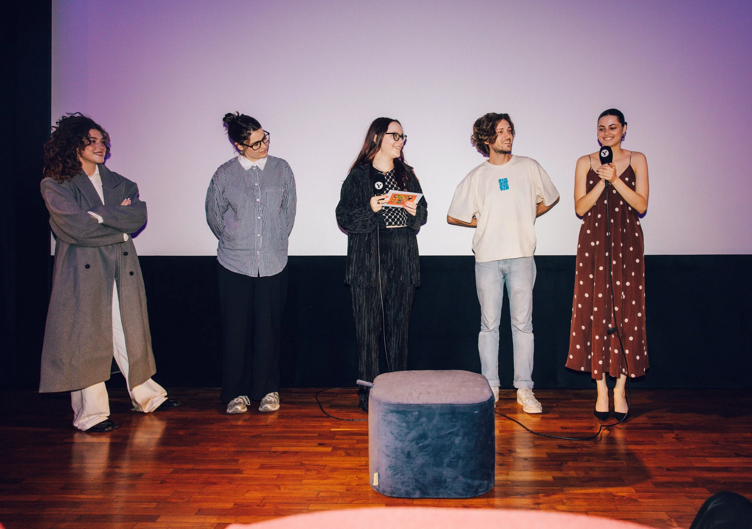 Five people stand on stage in front of a blank white screen, five women and one man, some holding microphones or papers, dressed in casual or semi-formal clothing, smiling, with a gray round stool on the wooden floor.