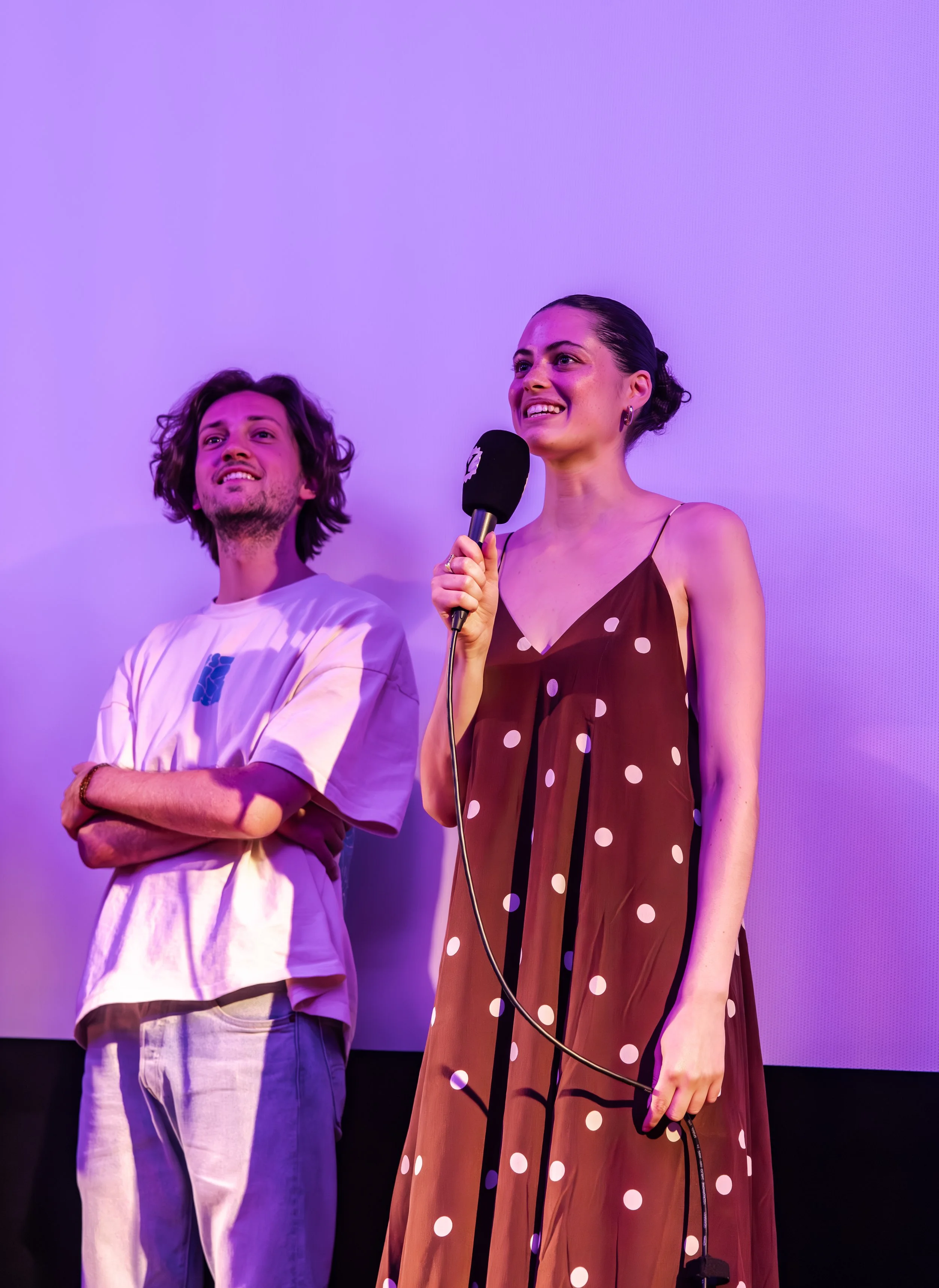 Two people on stage, one woman speaking into a microphone and one man standing with arms crossed, both smiling, against a purple background.