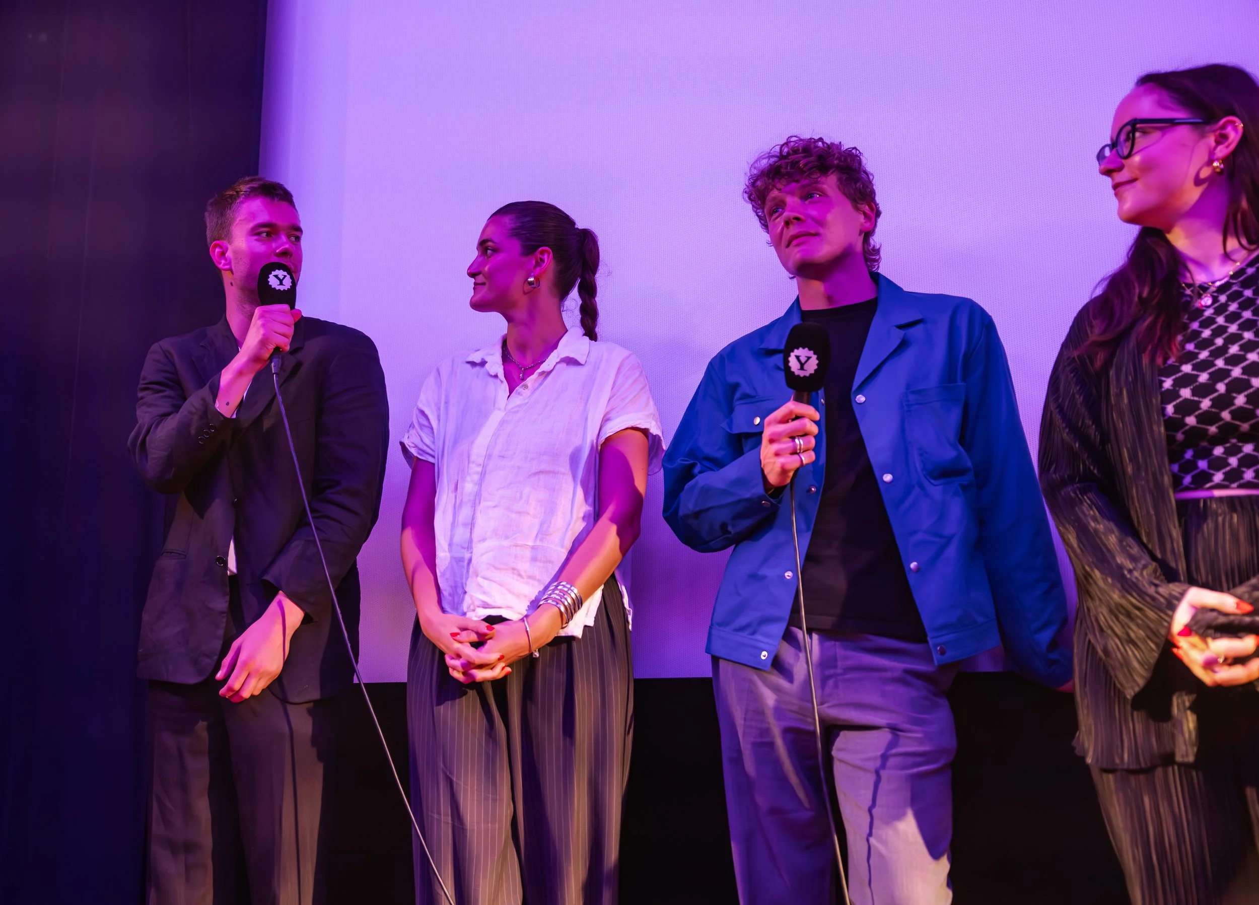 Four people standing on stage with microphones, likely at a panel or interview, illuminated by purple lighting.