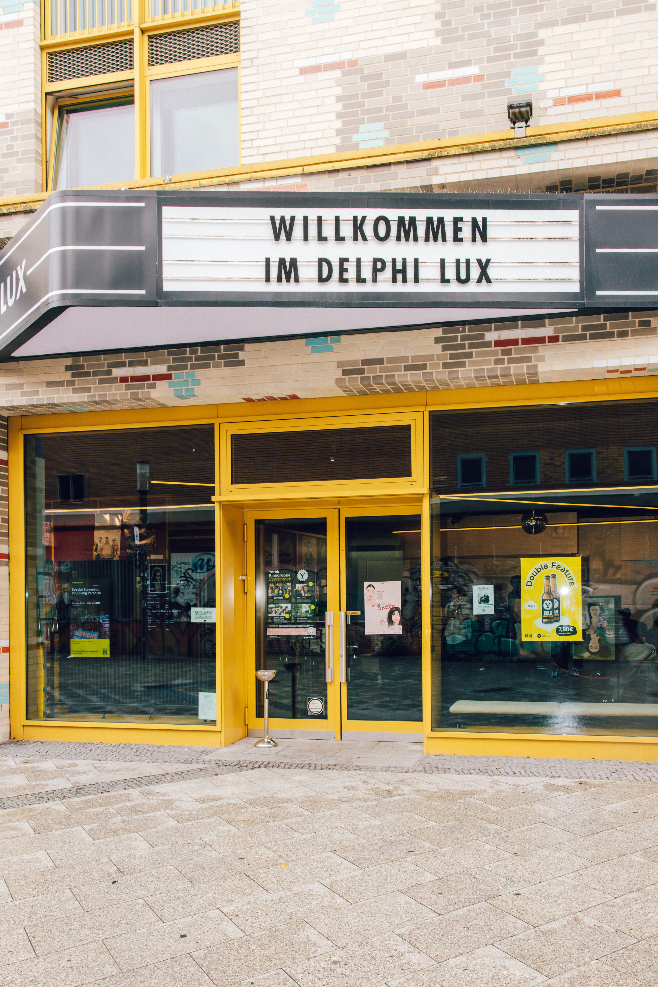 A storefront with yellow framing and large glass windows. Above, a marquee sign reads, "WILLKOMMEN IM DELPHI LUX". The sidewalk in front is paved with stone tiles.