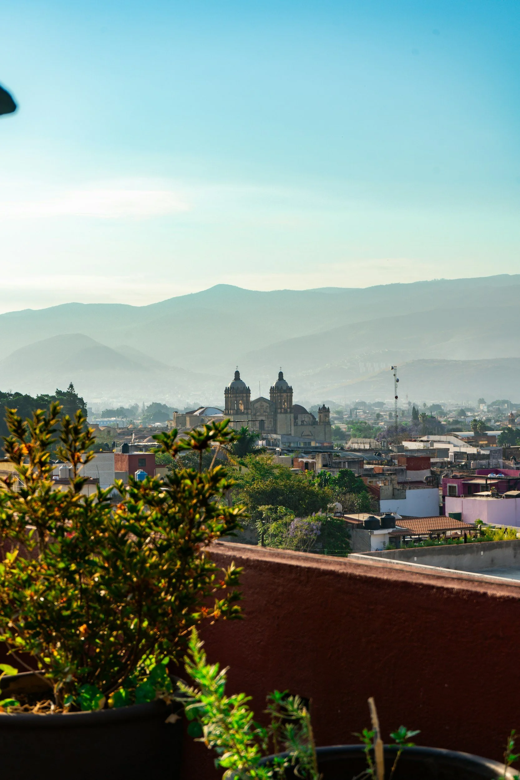 City skyline with a large historic church or cathedral with twin domes in the background, and mountains rising behind the city, taken from a balcony with potted plants in the foreground.