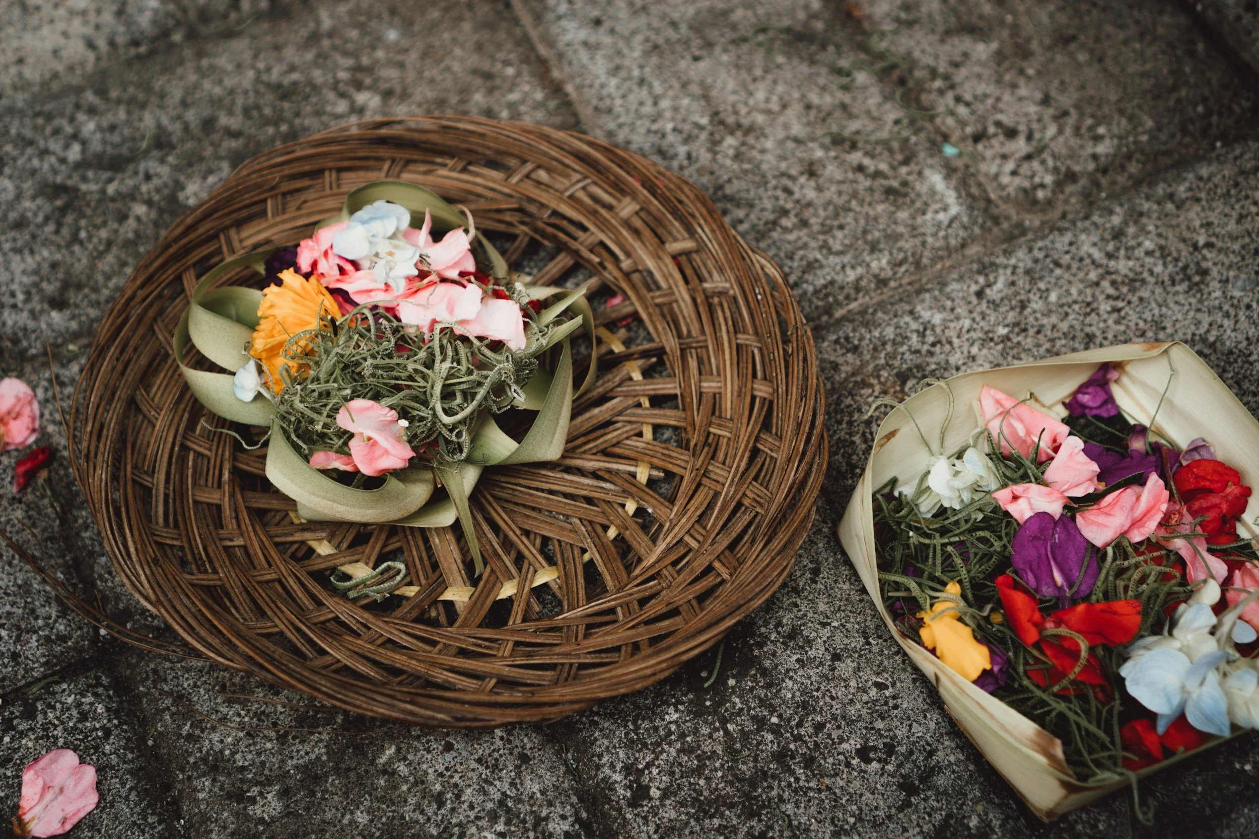 Colorful flower petals and greenery arranged on a woven basket and a square container, set on a stone surface.