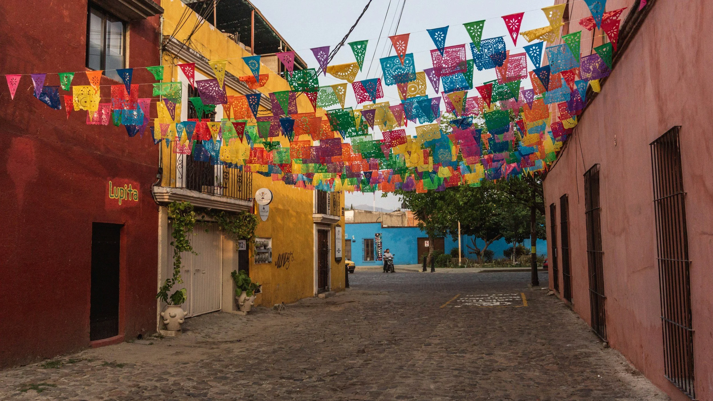 Colorful papel picado banners hang above a cobblestone street in a vibrant neighborhood with painted buildings in red, yellow, blue, and pink.