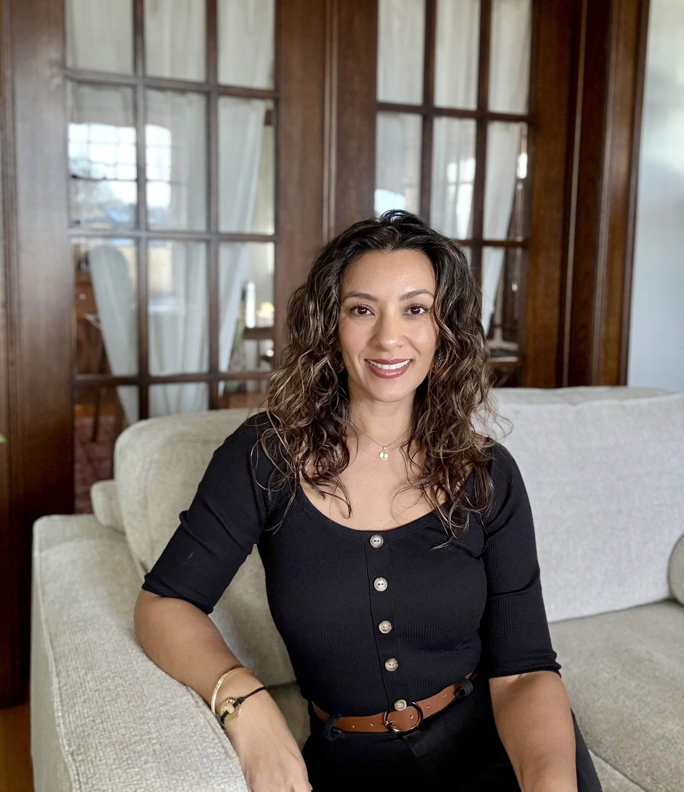 A woman with wavy brown hair smiling while sitting in a well-lit room with two black table lamps and clocks on the wall behind her.