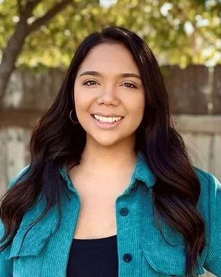 Young woman smiling outdoors with trees in the background, wearing a blue jacket over a dark top.