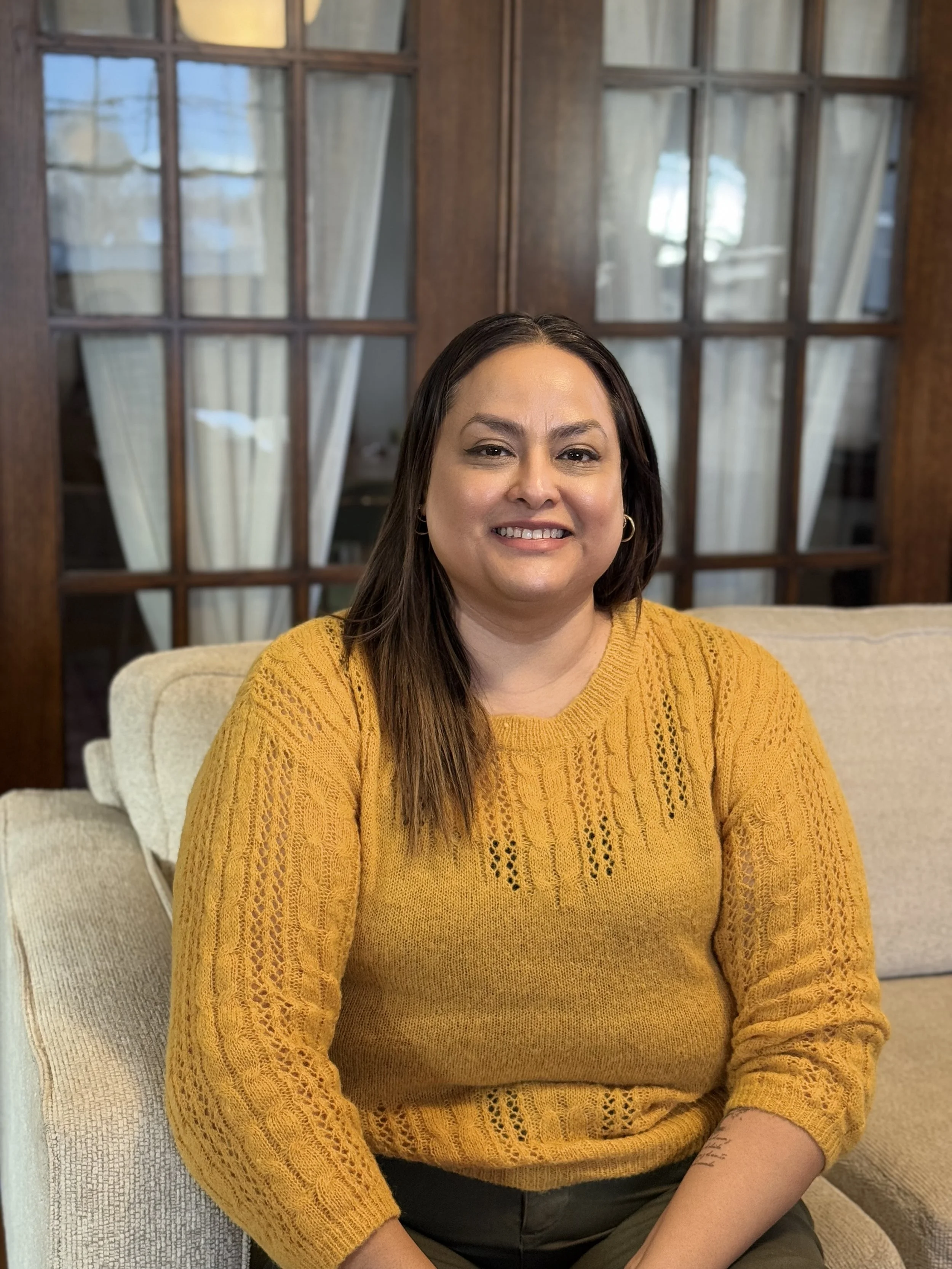 A woman sitting on a beige couch, smiling at the camera, in a cozy room with wooden furniture and green plants.