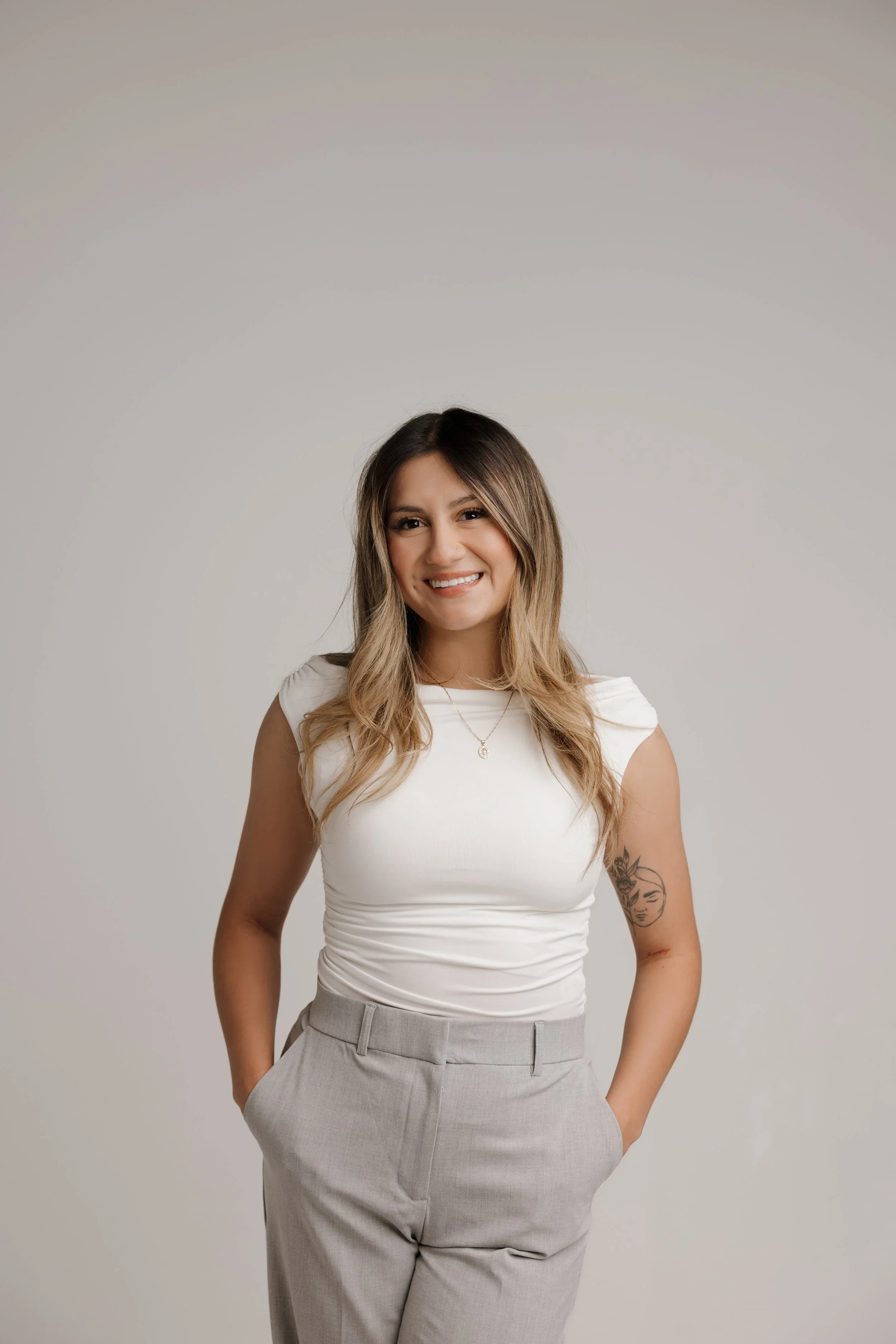 A woman with long, wavy hair wearing a white sleeveless top and light gray high-waisted trousers, smiling, standing against a plain light background.