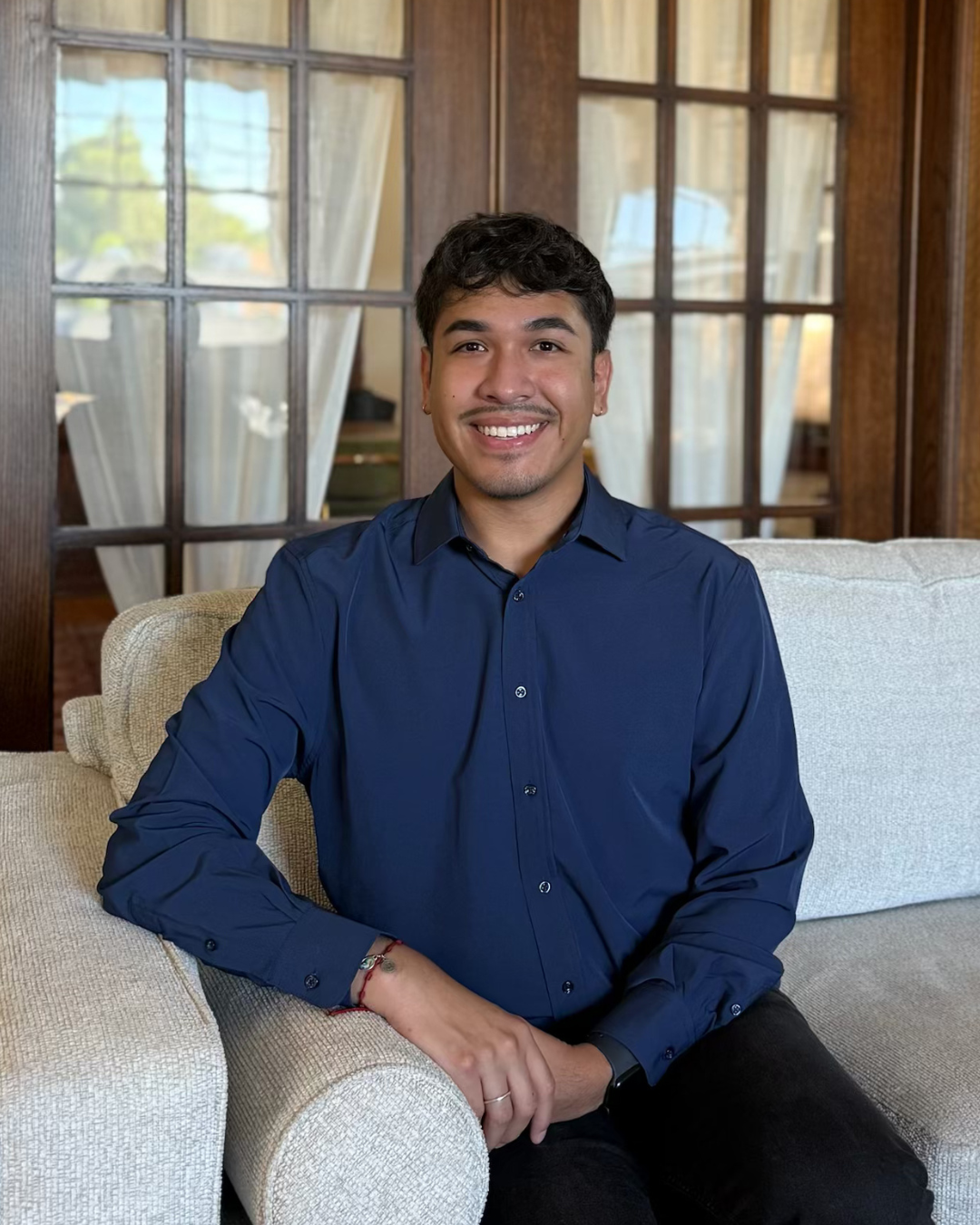A young man with dark hair and a mustache, wearing a navy blue button-up shirt, sitting on a beige sofa in a room with wooden walls and large windows with light curtains, smiling at the camera.