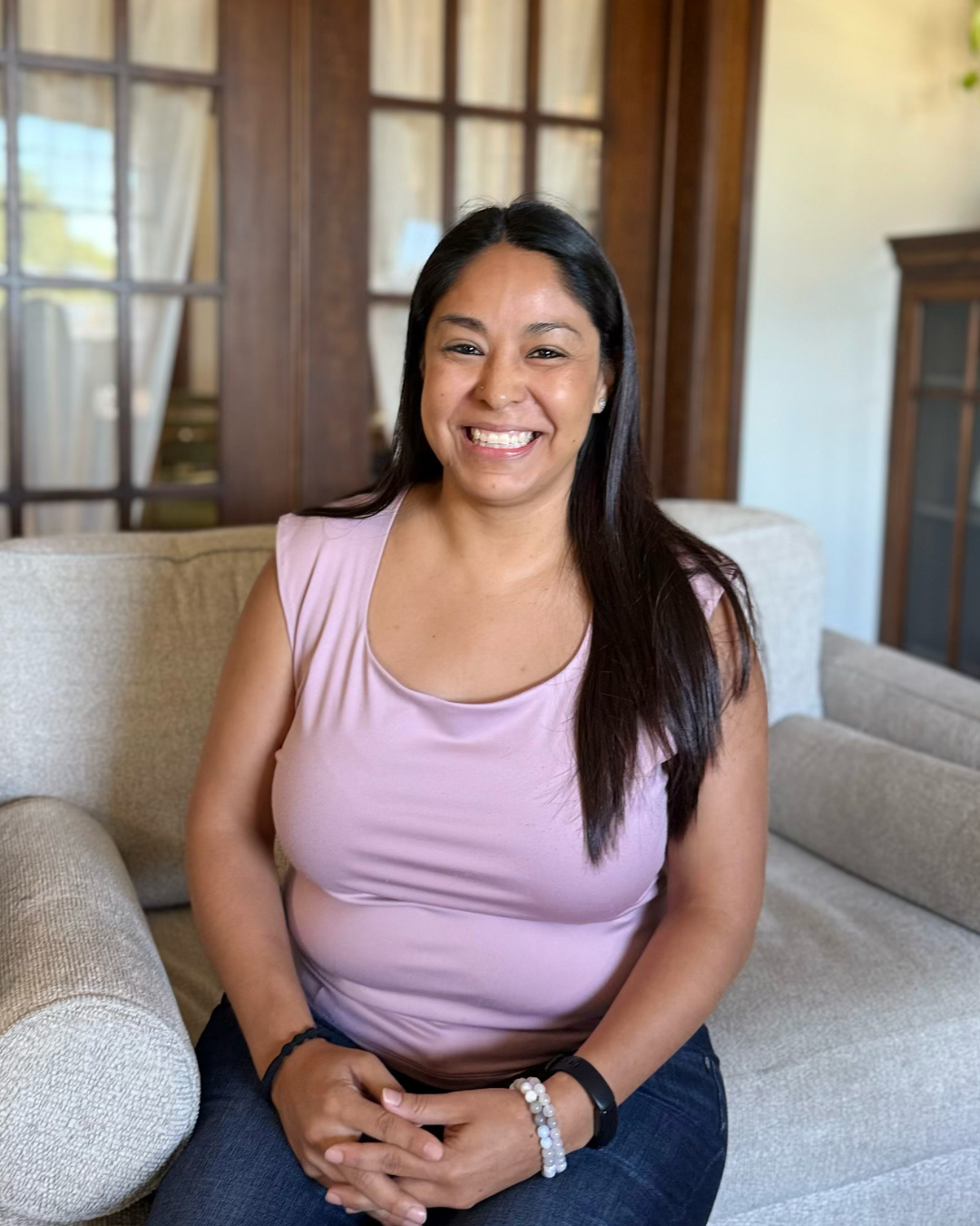 A woman with long dark hair, smiling, sitting on a beige sofa in a room with wooden walls and large glass-paneled doors in the background, wearing a pink sleeveless top and dark pants.