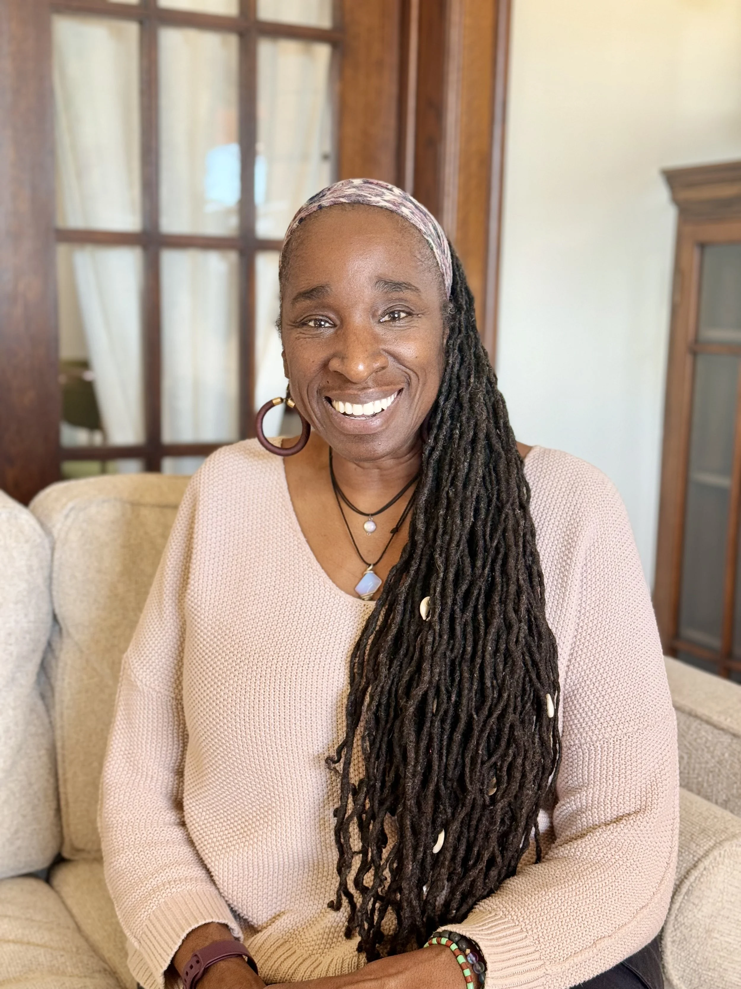 Smiling woman with long dreadlocks wearing an orange patterned dress, seated indoors, with a green plant in the background.