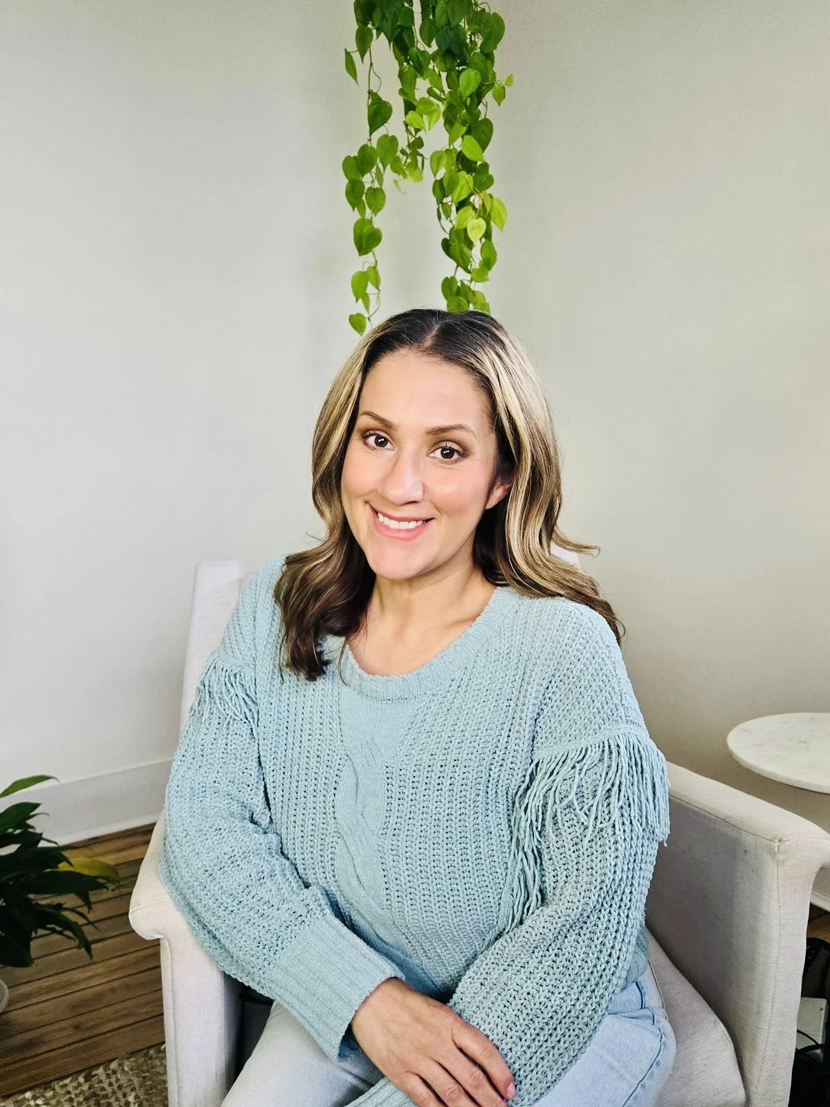 A woman with shoulder-length brown hair styled in loose waves, wearing makeup and red earrings, smiling while standing outdoors next to a tree, dressed in a burnt orange off-the-shoulder top with spaghetti straps.