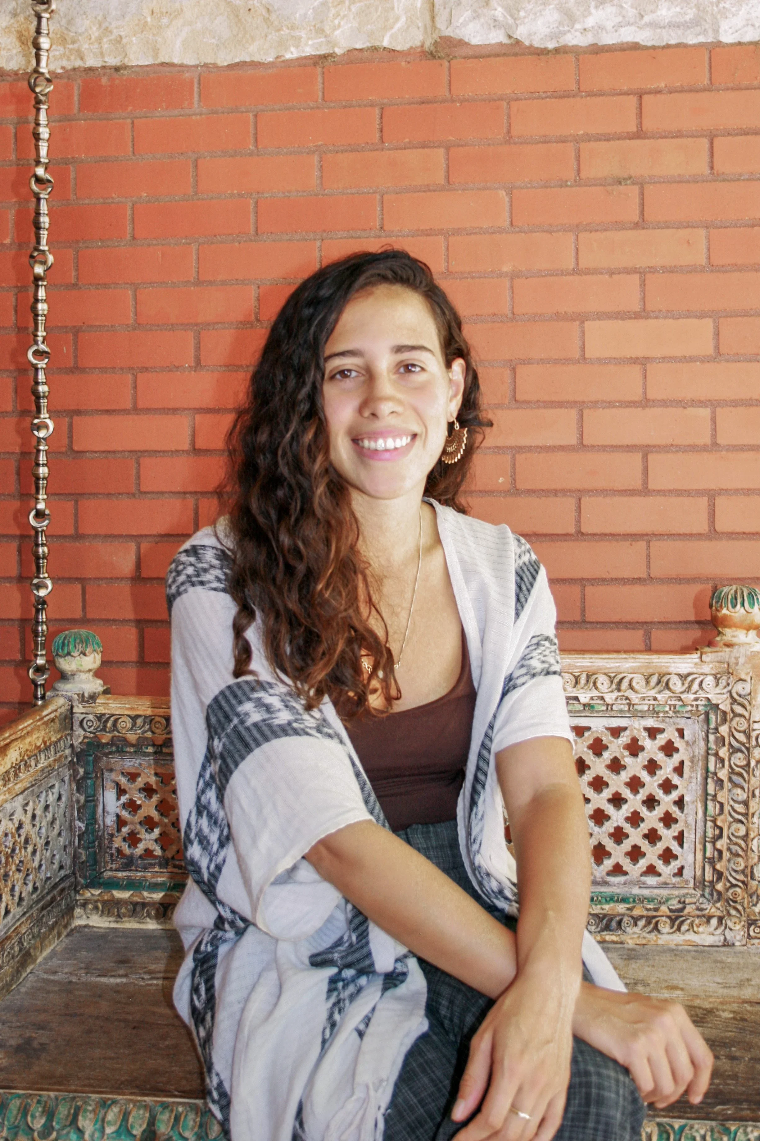A woman with long, curly brown hair, wearing a brown top and a white and black patterned shawl, sitting on an ornate, wooden bench with a red brick wall behind her, smiling at the camera.