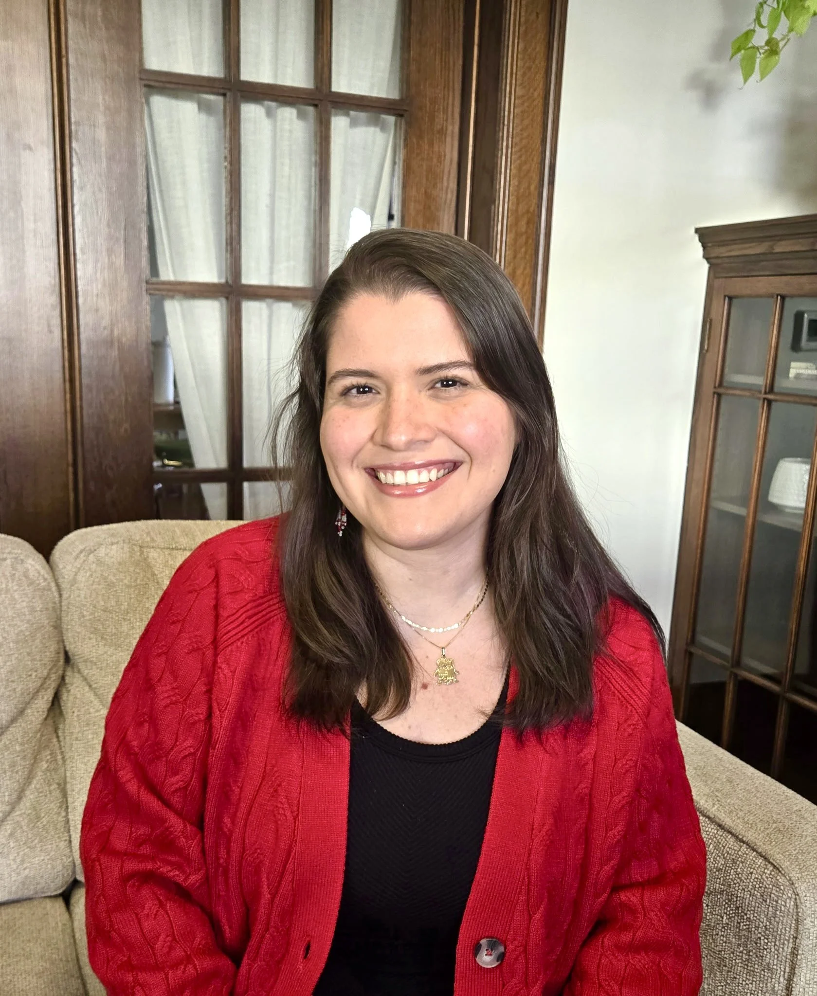 Woman with shoulder-length dark hair and a pink streak, wearing a patterned blazer and a bright pink top, smiling in front of a brick wall.